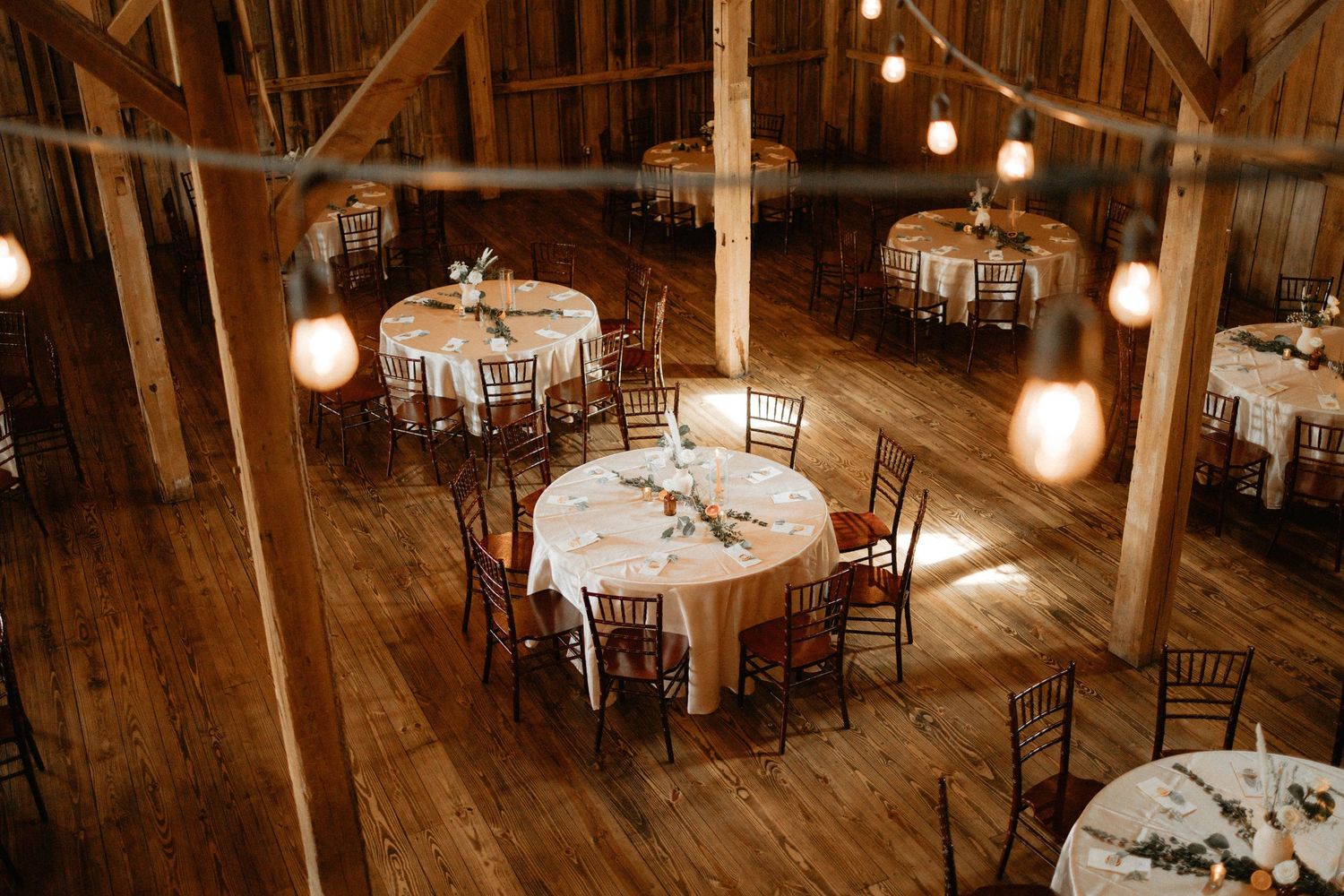 Rustic barn wedding reception setup with round tables, wooden chairs, and hanging lights on exposed wooden beams.