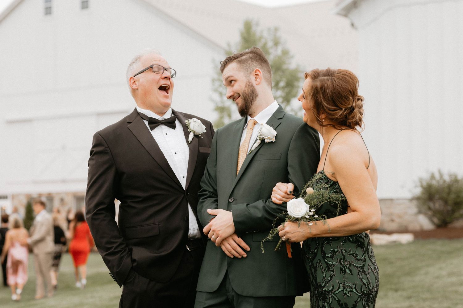 Wedding party members share joyful candid moments on a grassy lawn next to a white barn building.