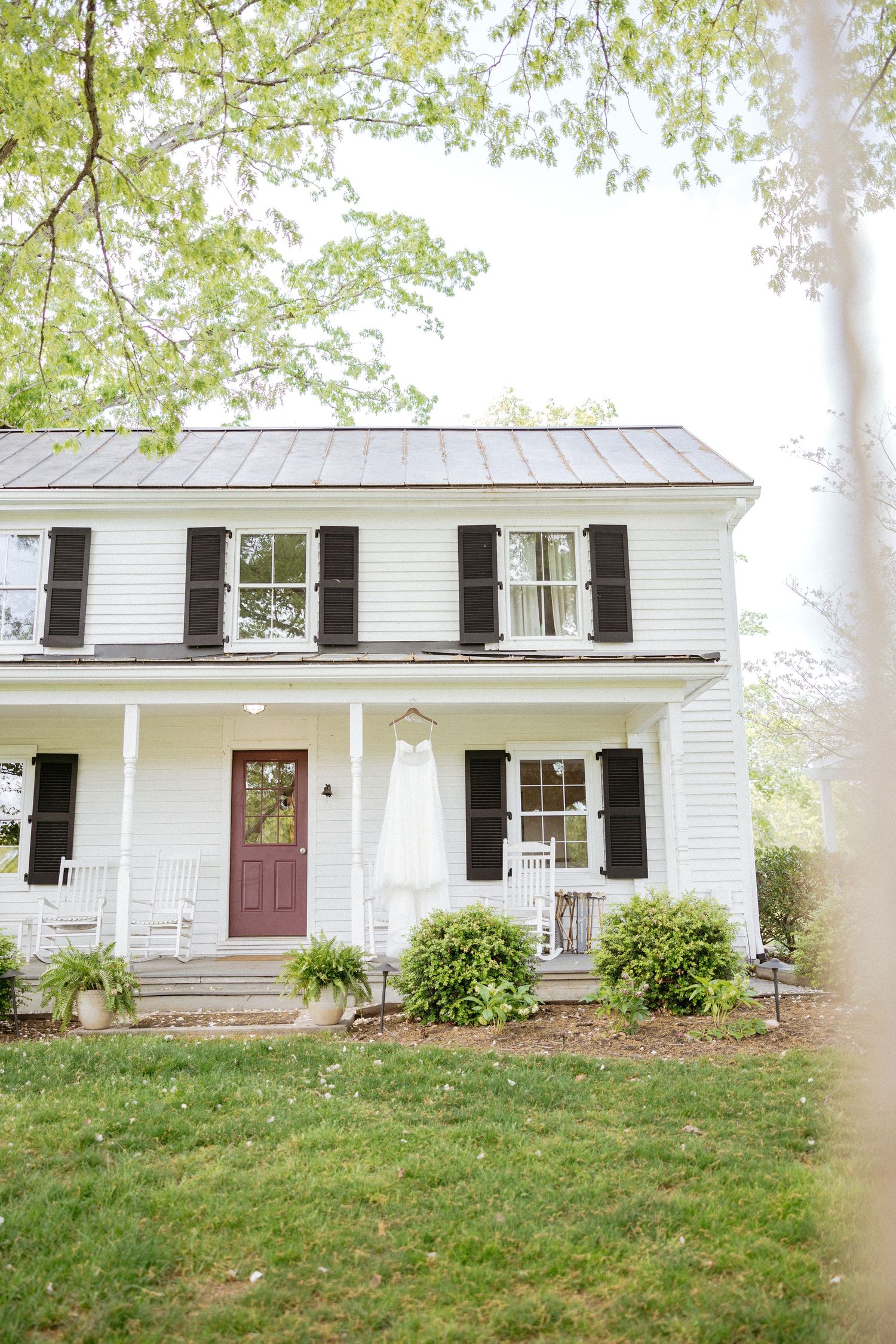 Charming white farmhouse with black shutters features a welcoming front porch adorned with greenery and a red door.