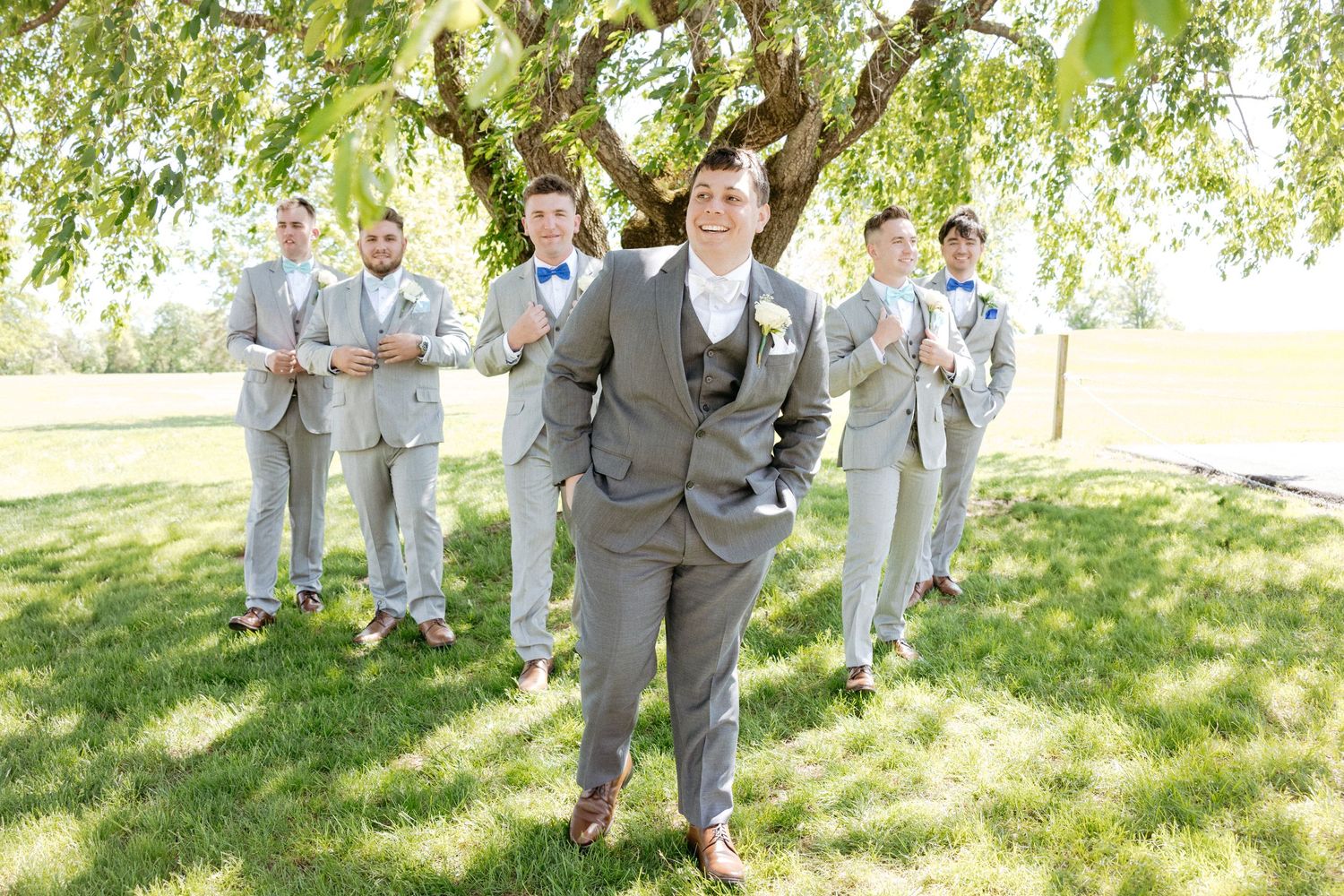 Groomsmen in light grey suits pose outdoors under a willow tree.