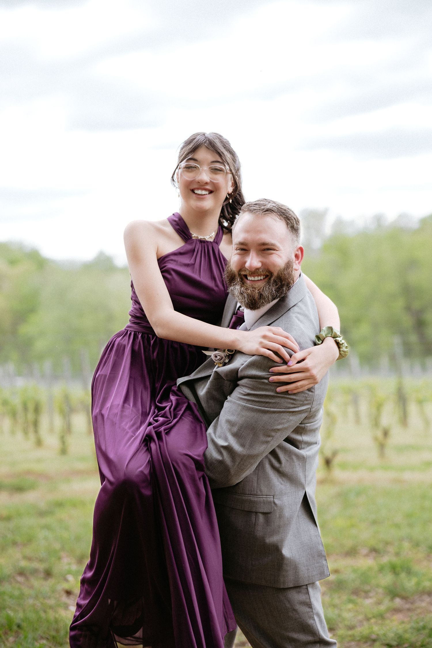 A couple embraces and laughs together in a field wearing a purple dress and grey suit.
