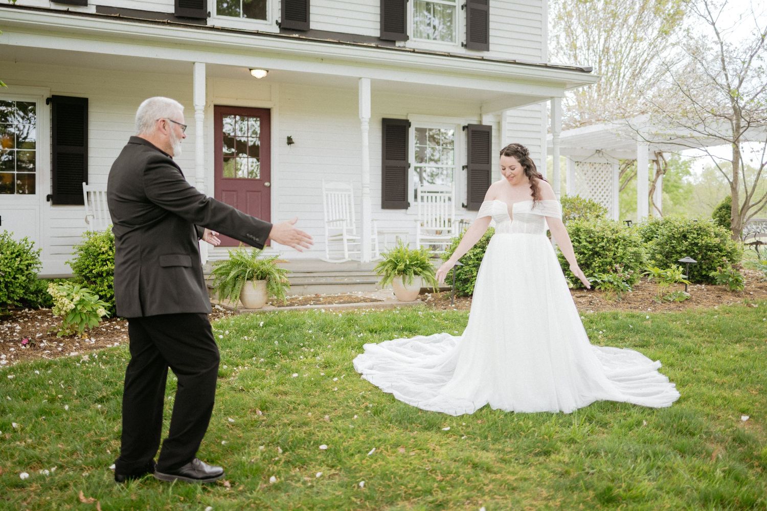 Wedding party poses for outdoor photos on a grassy lawn in front of a white house with a wrap-around porch.