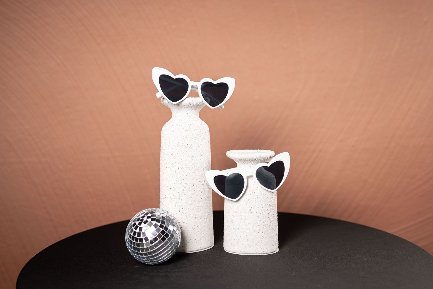 Black heart sunglasses and Disney-themed items arranged with white objects and disco ball.