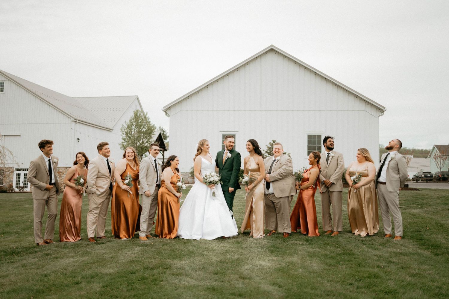 Wedding party members in orange and beige attire pose in front of a white barn for group photos.
