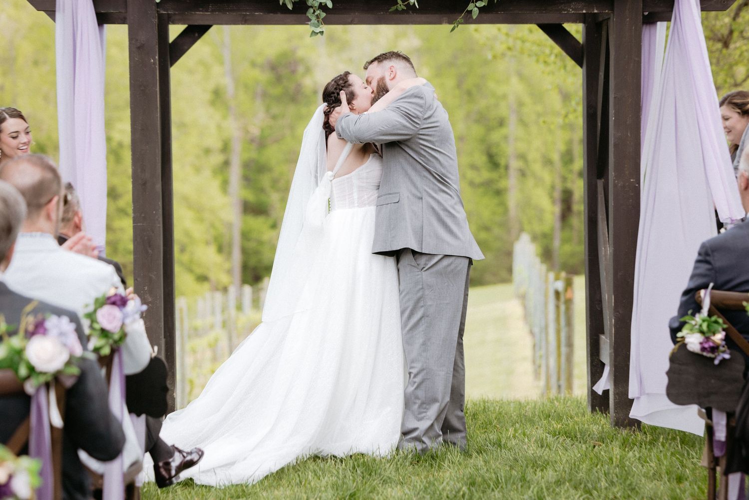 A romantic outdoor wedding ceremony with couples embracing under a white fabric draped arch amidst green trees.