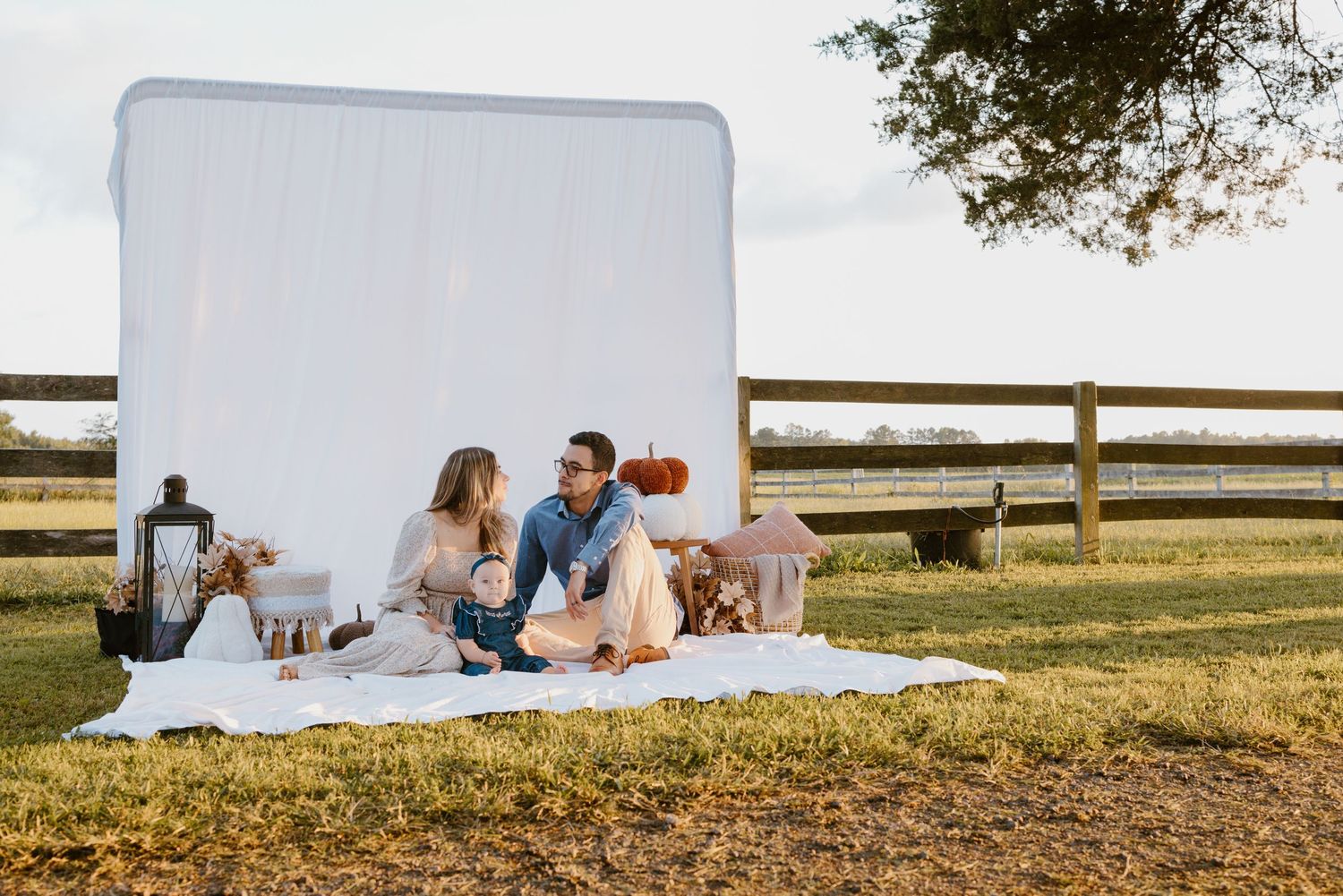 A family enjoys picnicking together on a white blanket near a rustic wooden fence during sunset.