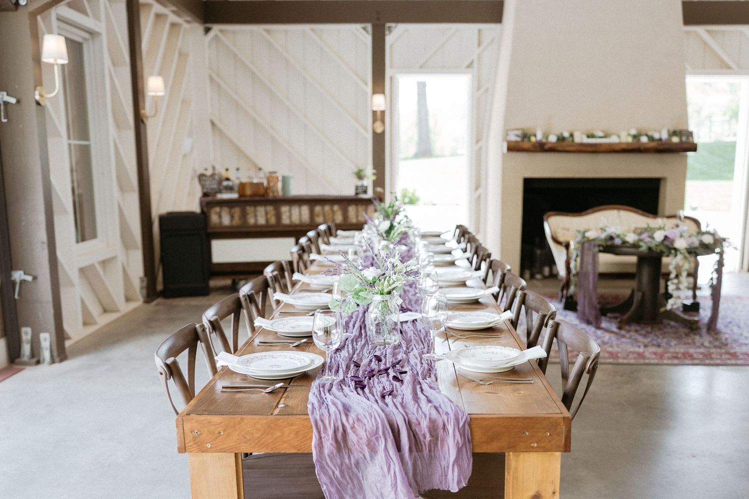 A long wooden dining table set with white plates and a flowing purple table runner in a rustic white-walled venue.