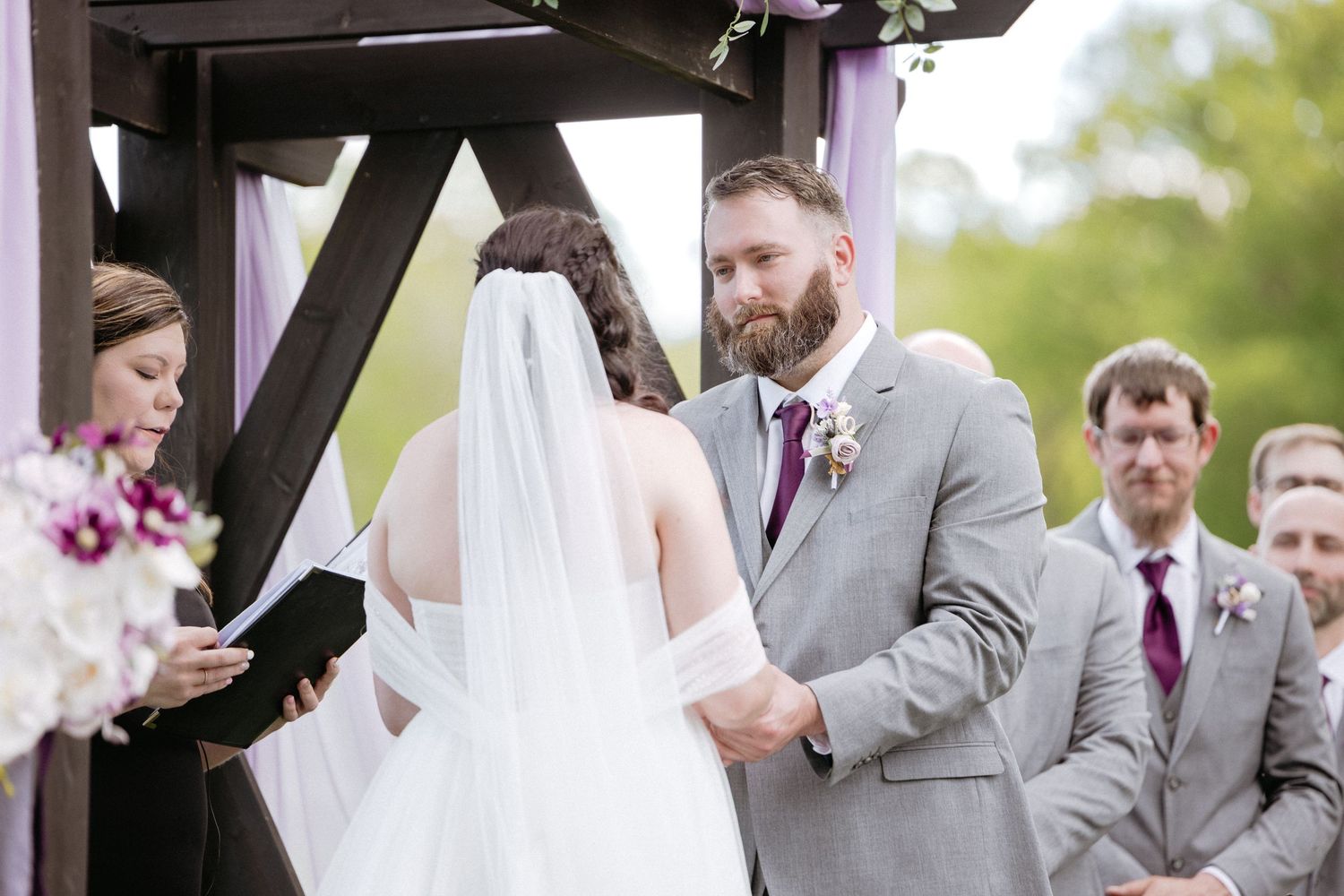 Wedding party gathered under wooden gazebo during outdoor ceremony with purple floral decor.