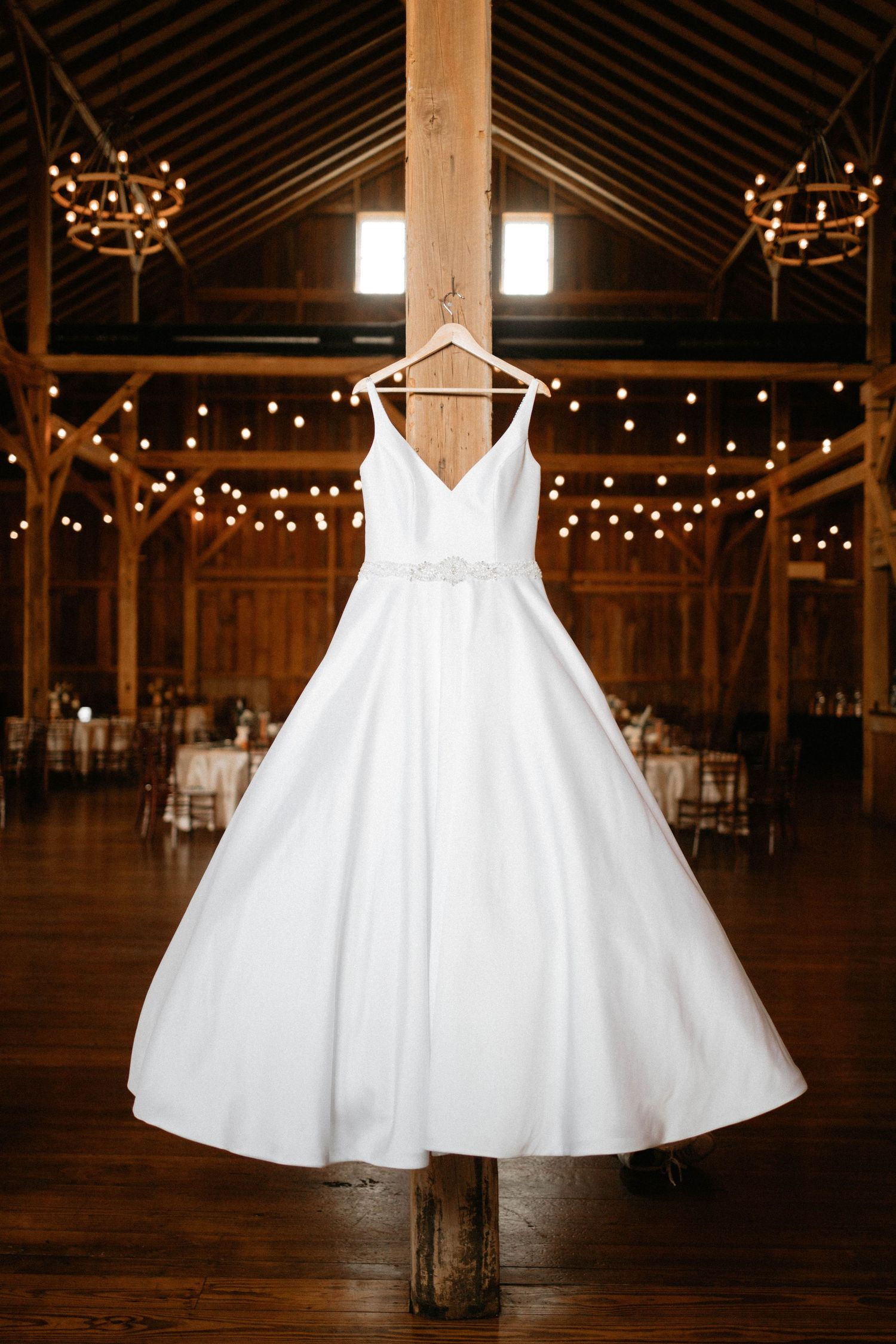 A white wedding dress hangs elegantly in a rustic barn venue decorated with twinkling string lights.