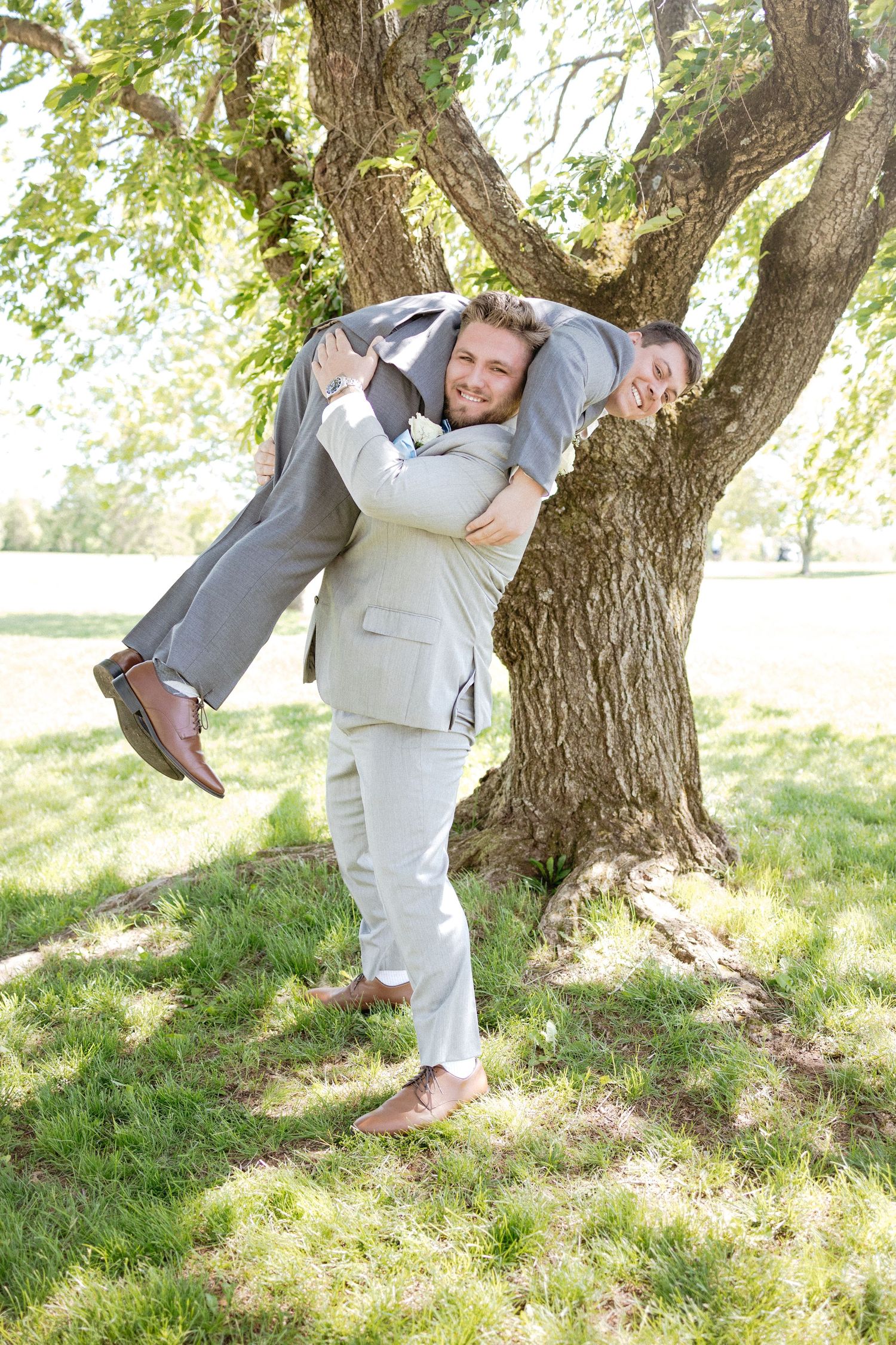 Playful wedding photos capture intimate moments between a couple near a large tree on a grassy lawn.