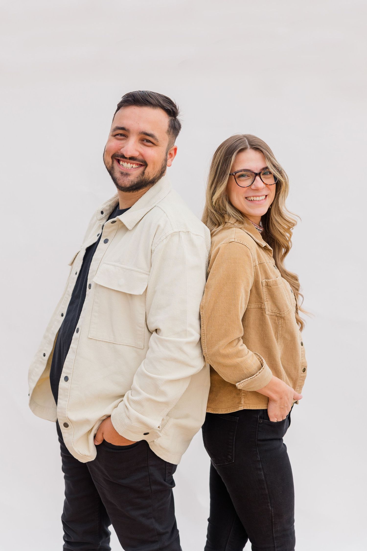 Two people dressed in contrasting black and beige outfits pose together for stylish studio portraits on white background.