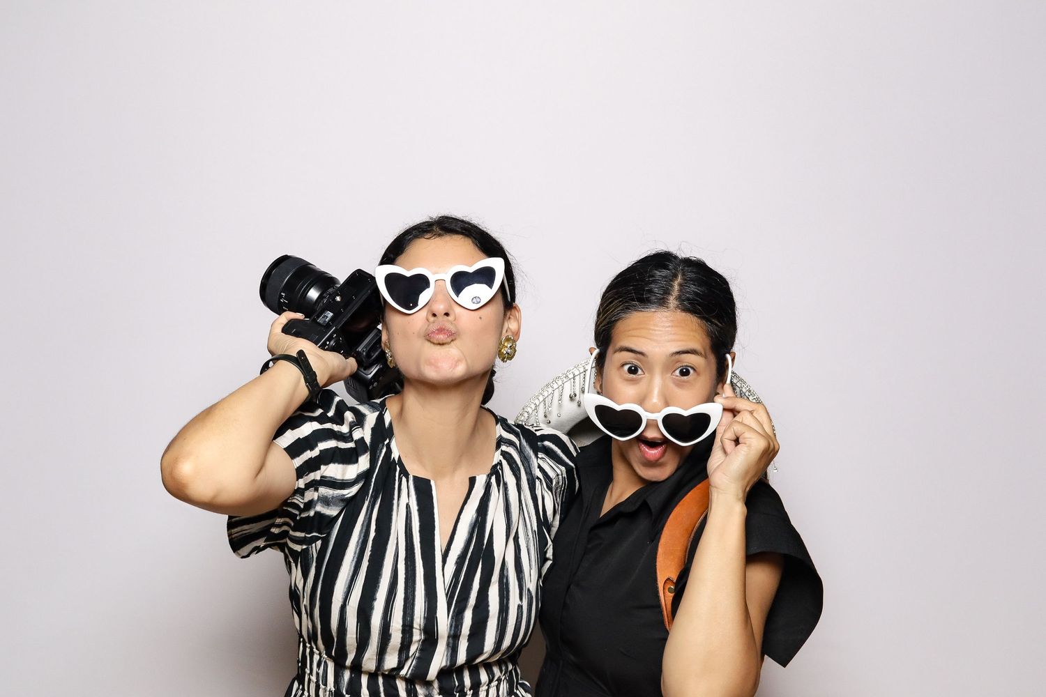 Two people in black and white outfits wearing sunglasses pose playfully with a camera against a light gray background.