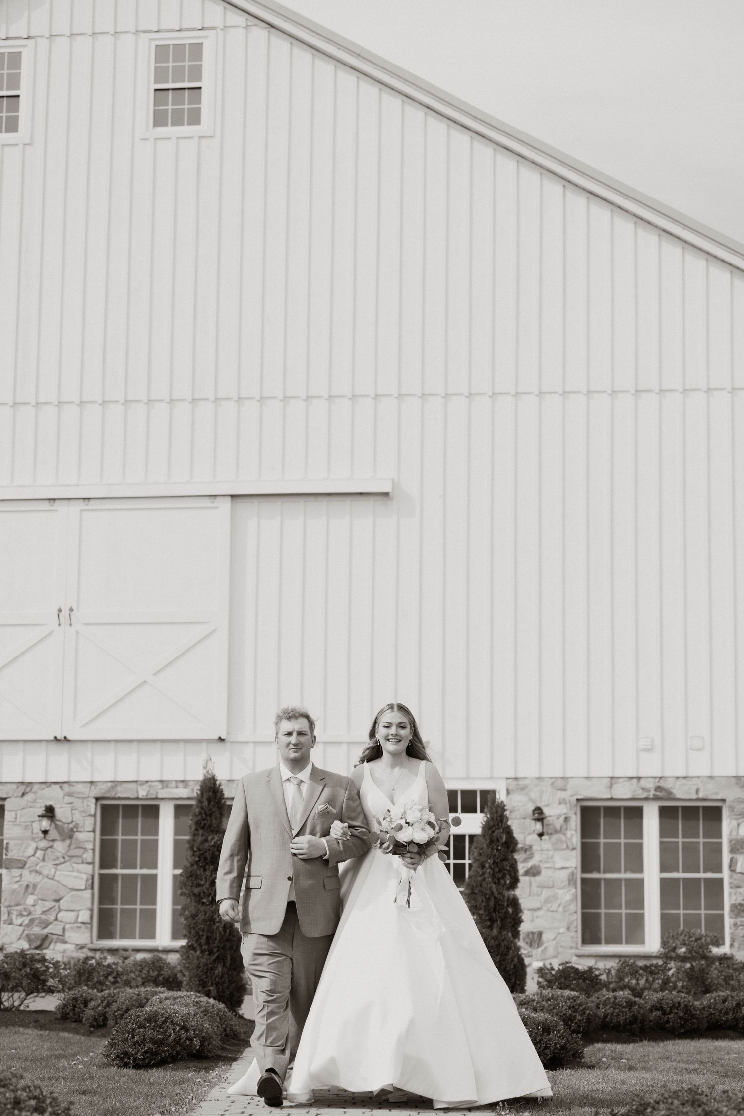 A wedding couple poses in front of a classic white barn building with stone foundation.