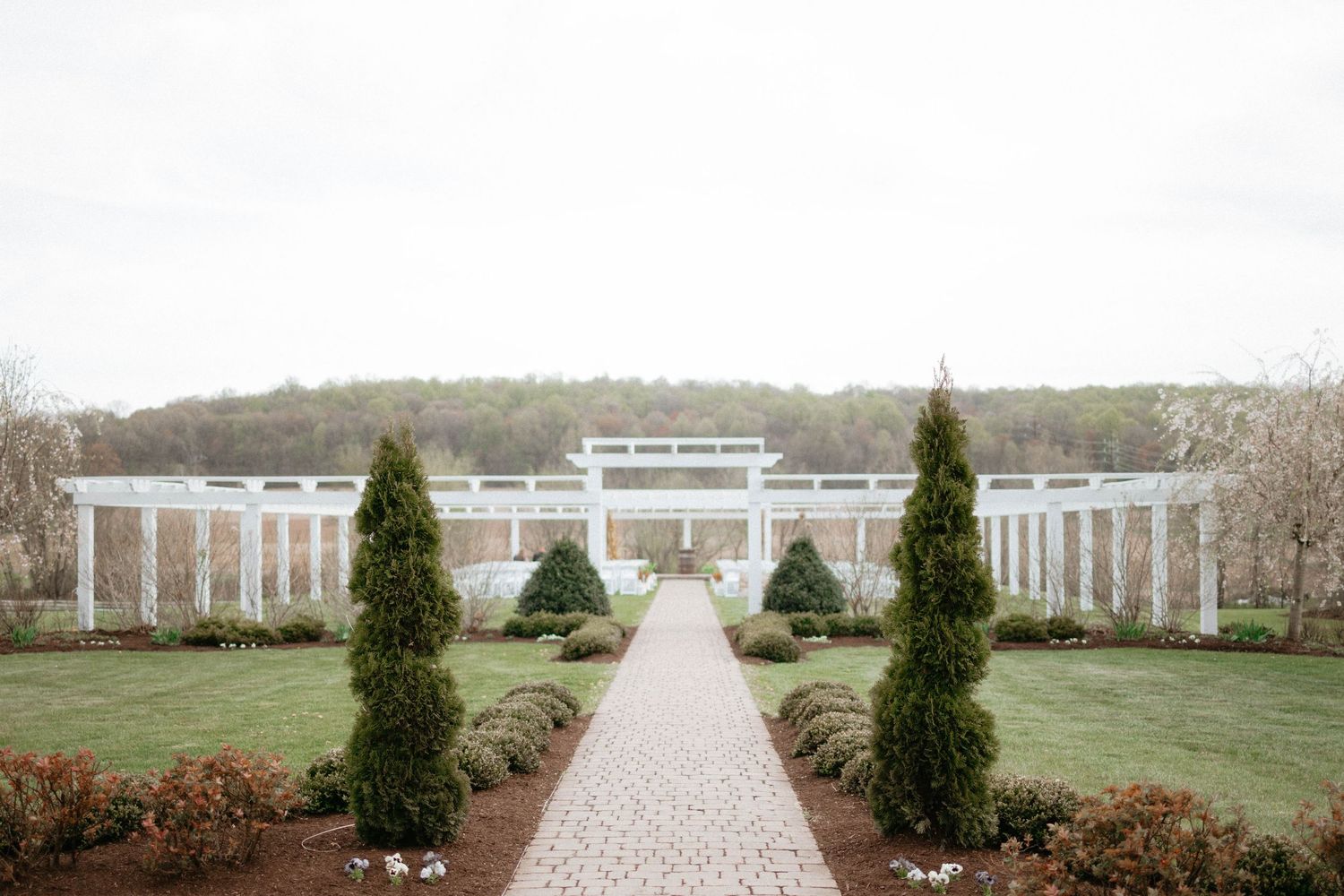 A curved garden pathway lined with evergreen trees leads to a white colonial-style building in the distance.