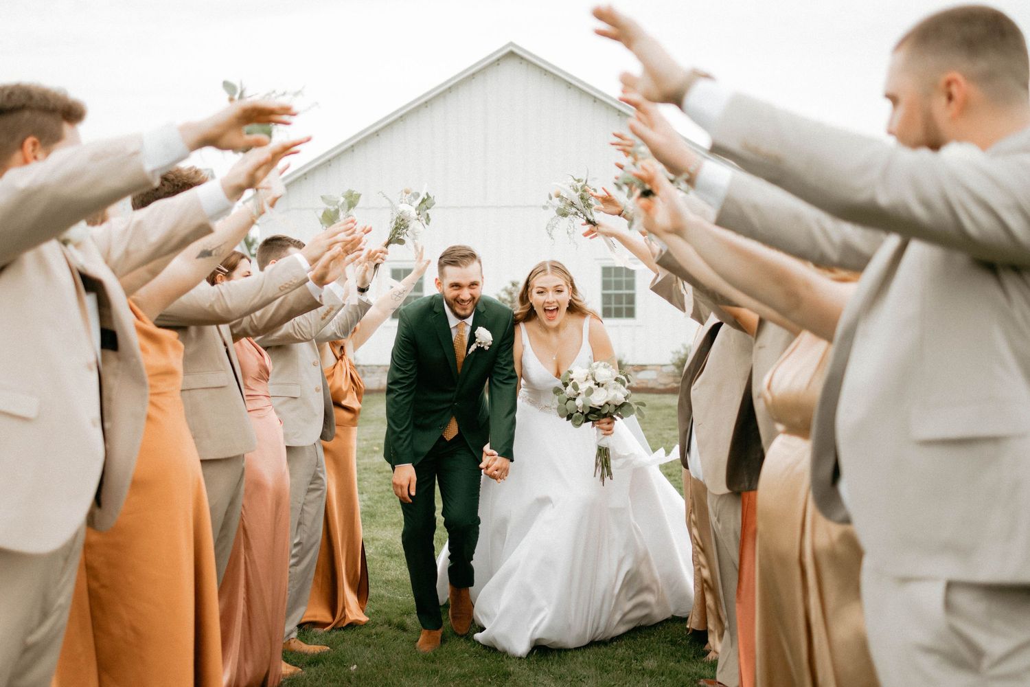 Wedding party members celebrate with raised hands in front of a rustic white barn.