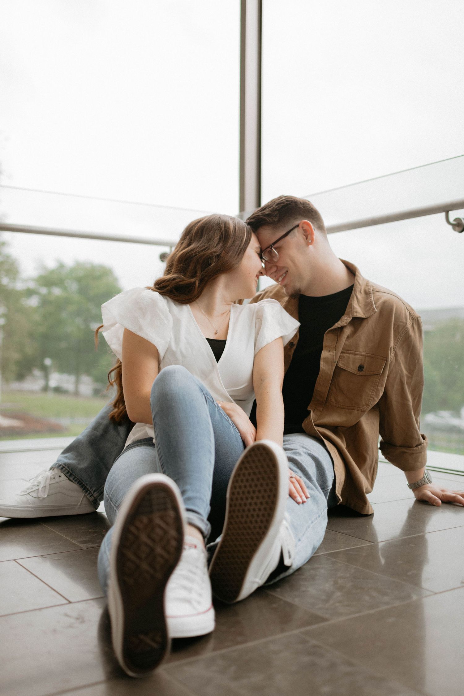 Couple sitting on floor together in casual outfits sharing intimate moment by window during indoor photoshoot.