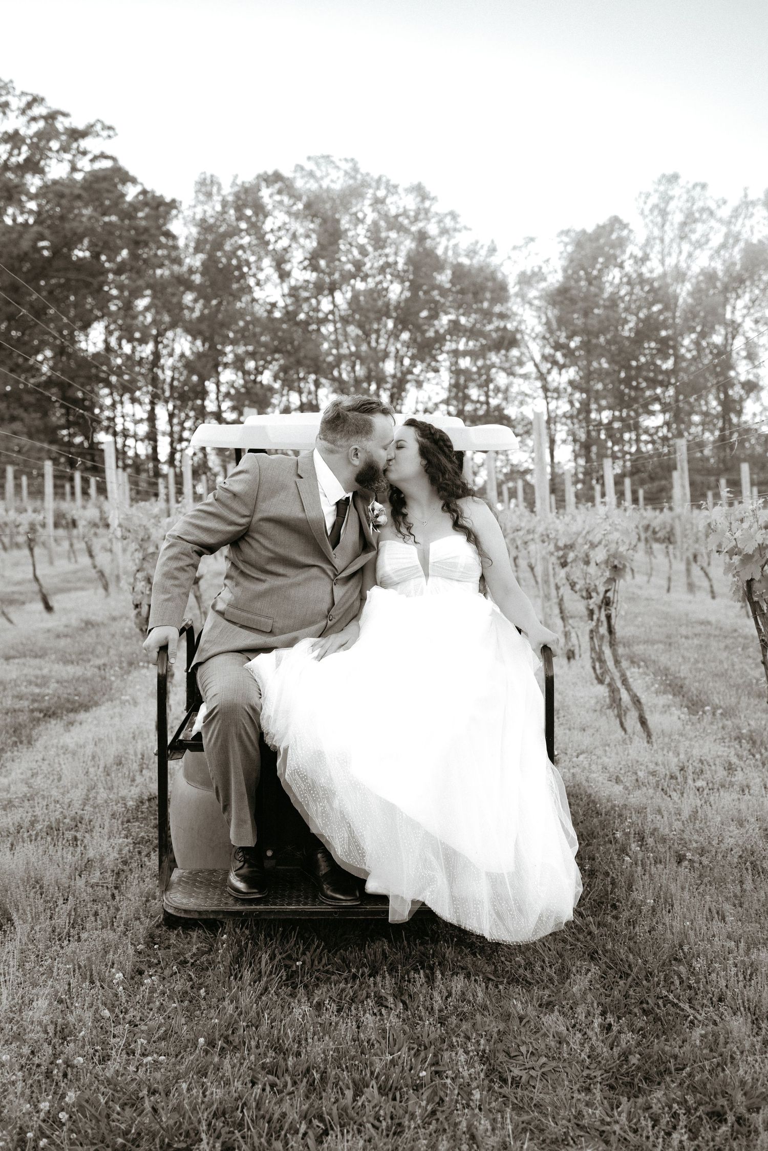 A tender romantic moment captured in black and white as a couple sits together in a grassy field with trees in the background.