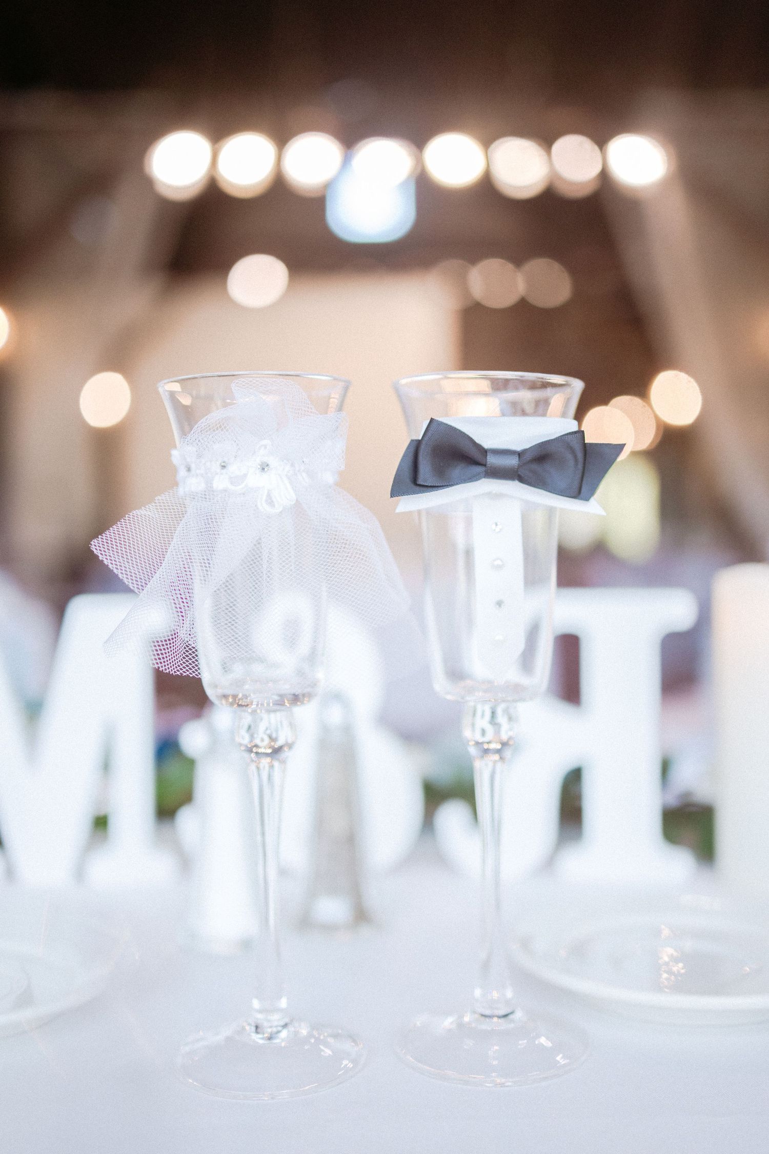 Wedding champagne glasses decorated with elegant bride and groom accessories against bokeh light background.