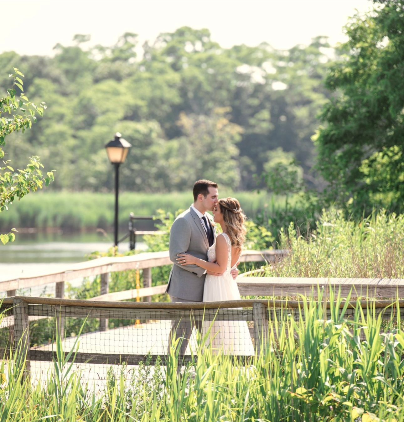 A romantic couple shares a tender kiss on a wooden boardwalk surrounded by lush summer greenery and wetlands.