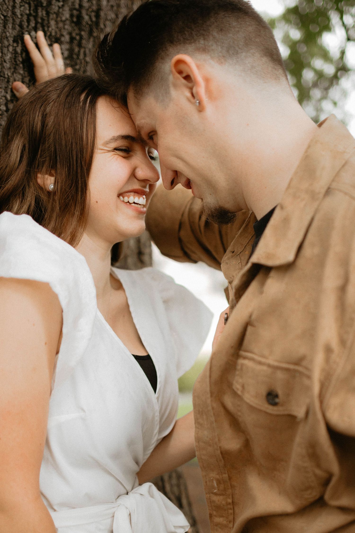 Romantic couple in white and tan clothing share an intimate moment outdoors with foreheads touching and joyful smiles.