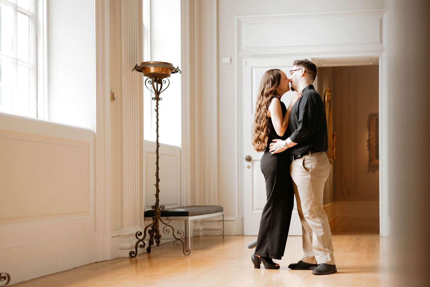 A couple in elegant formal wear dances together in an elegant white room with tall windows and natural light.