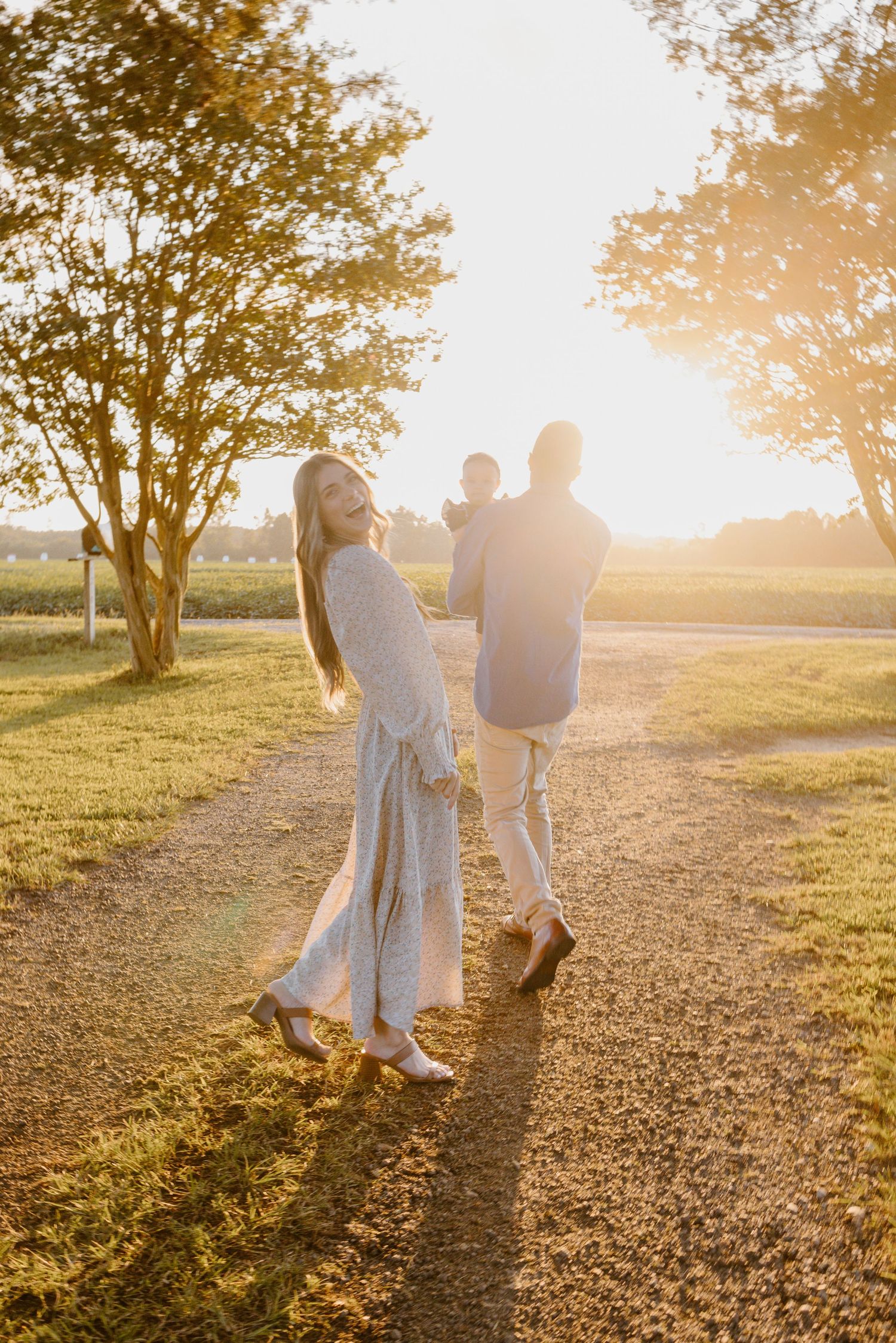 A couple walks hand in hand during golden hour sunset casting long shadows across a grassy field beneath trees.