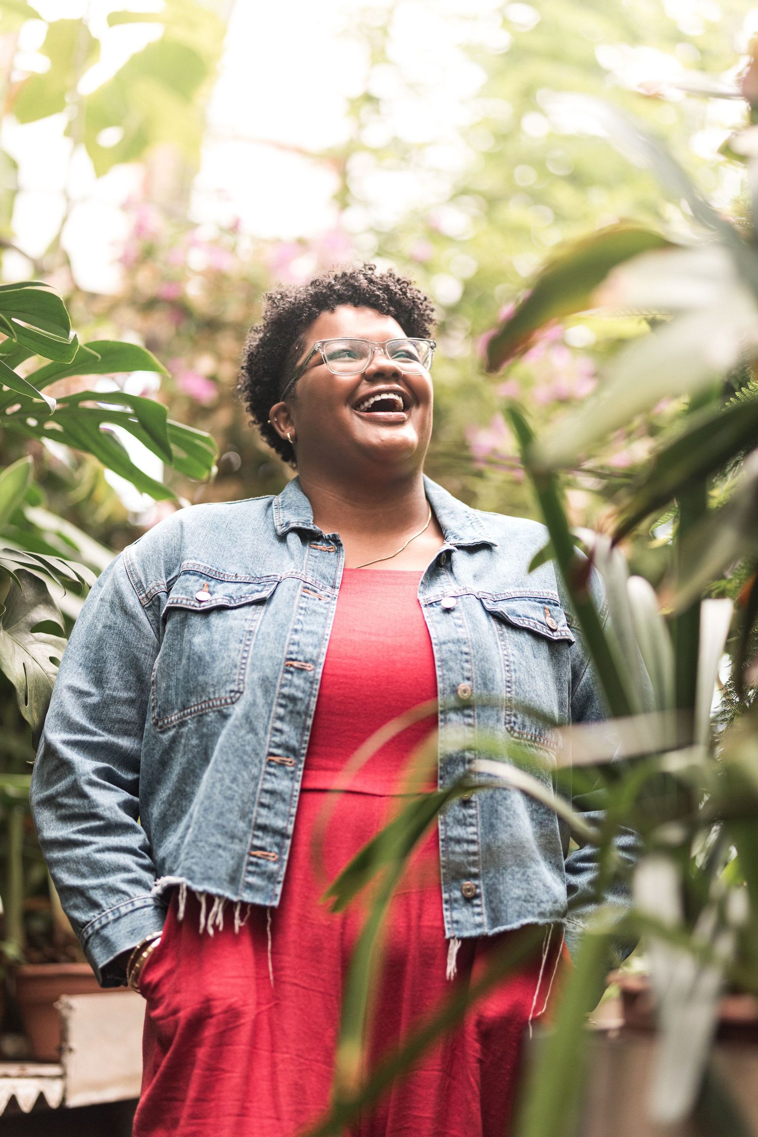 A person in a denim jacket and red shirt smiles joyfully while standing among lush green plants in a garden setting.