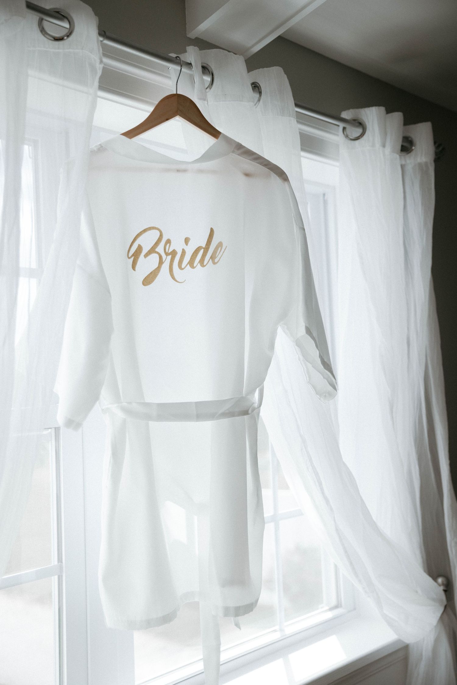 White bridal robe with gold 'Bride' text hanging on wooden hanger against sheer curtains.