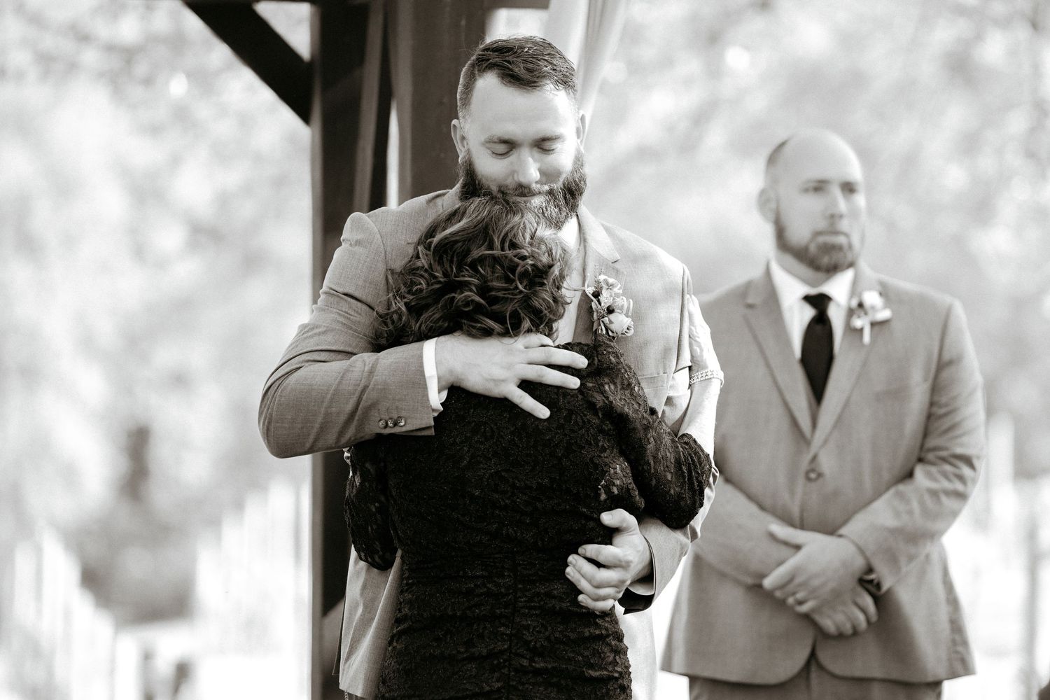 Two people share an emotional embrace during a wedding ceremony while a suited guest looks on in the background.