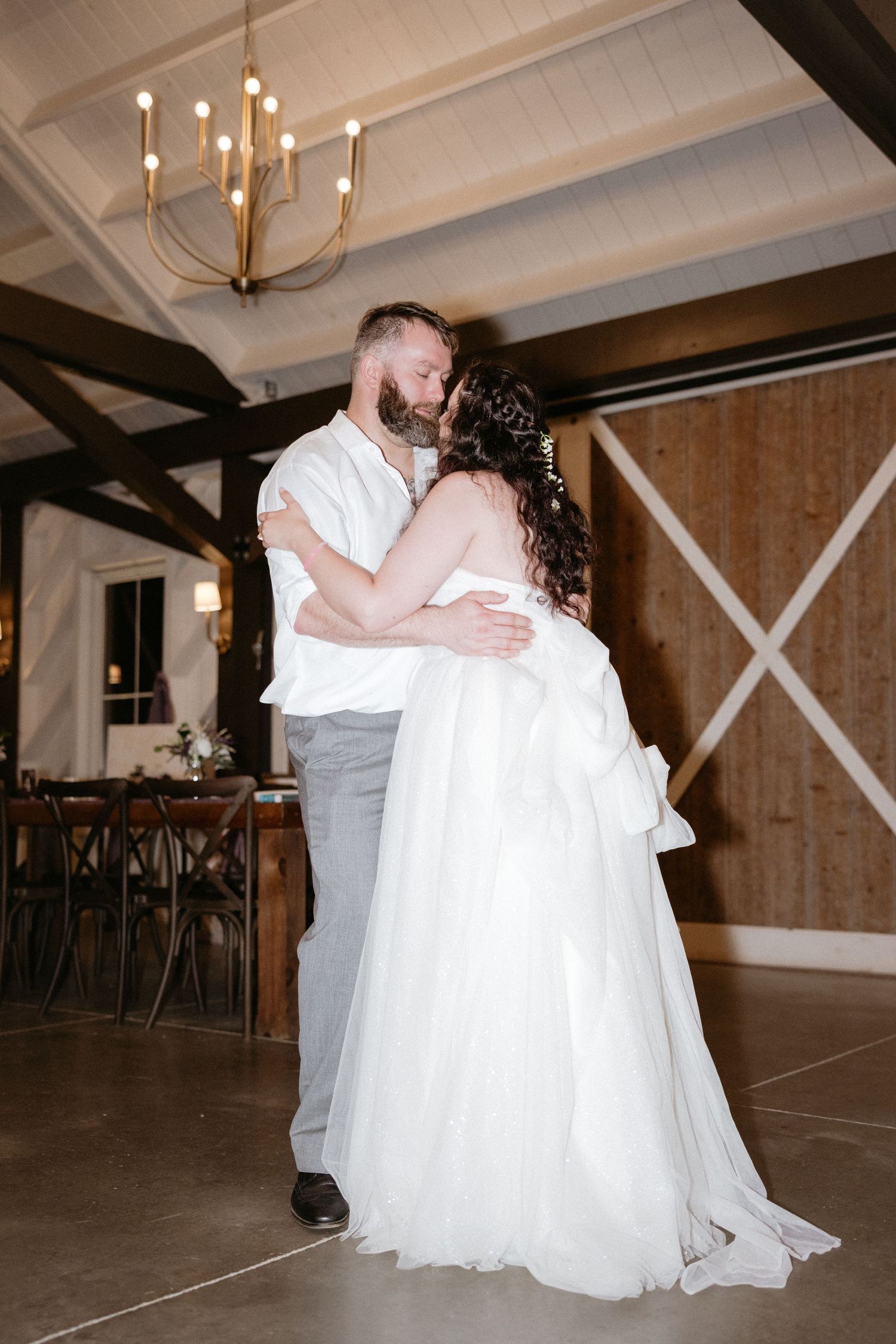 Couple shares first dance in rustic barn venue with wooden beams and chandelier lighting.