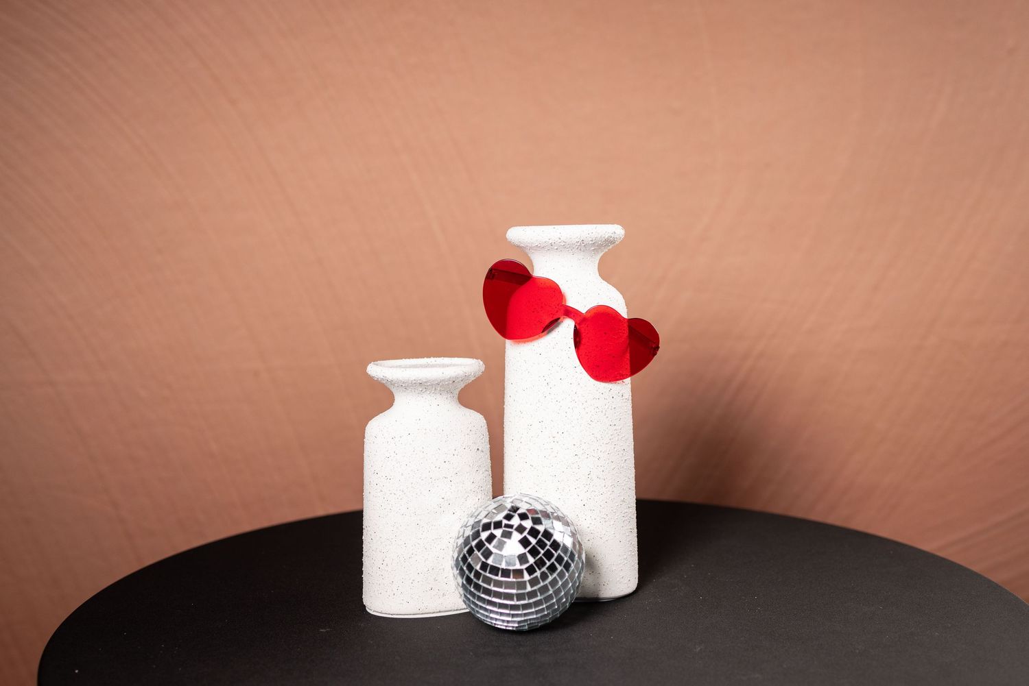 Red heart-shaped sunglasses arranged with white objects and silver disco ball on brown background.