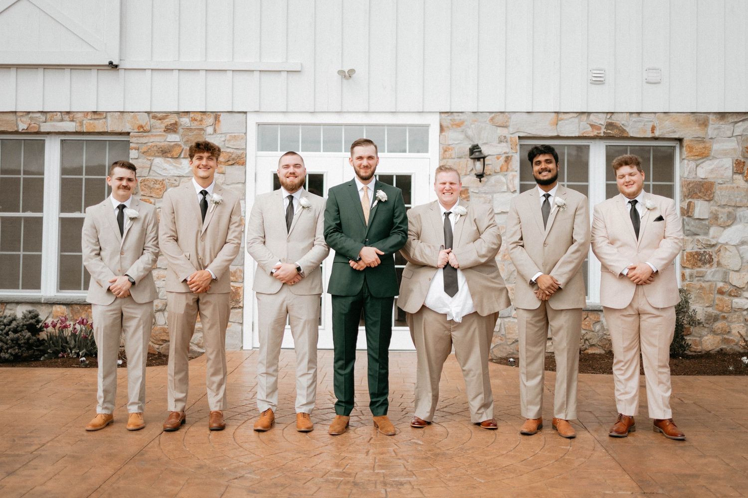 Wedding party poses against stone wall exterior wearing coordinated beige suits and dark green accents.