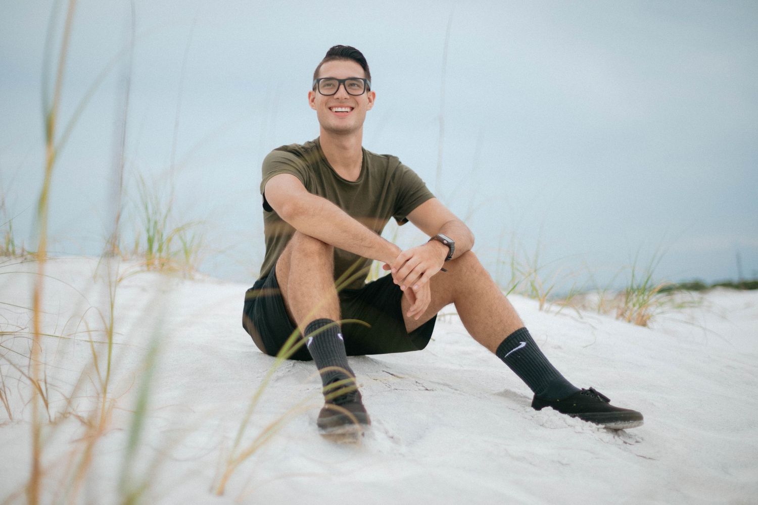Someone sits casually on white sand beach while wearing glasses, shorts and dark t-shirt.