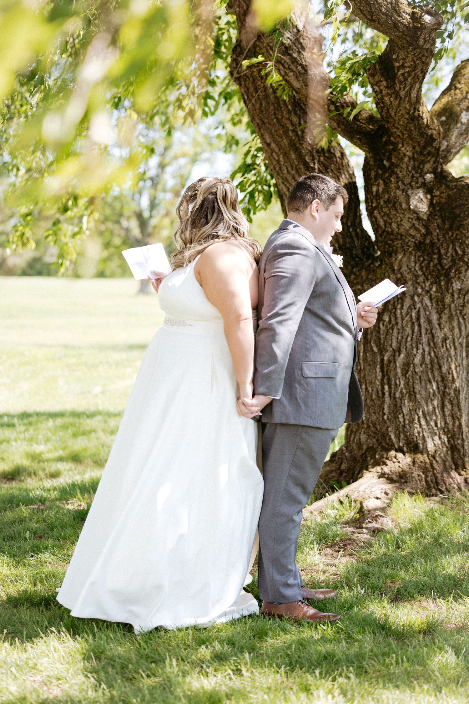 Wedding couple stands near a tree in a sun-dappled orchard setting on their special day.