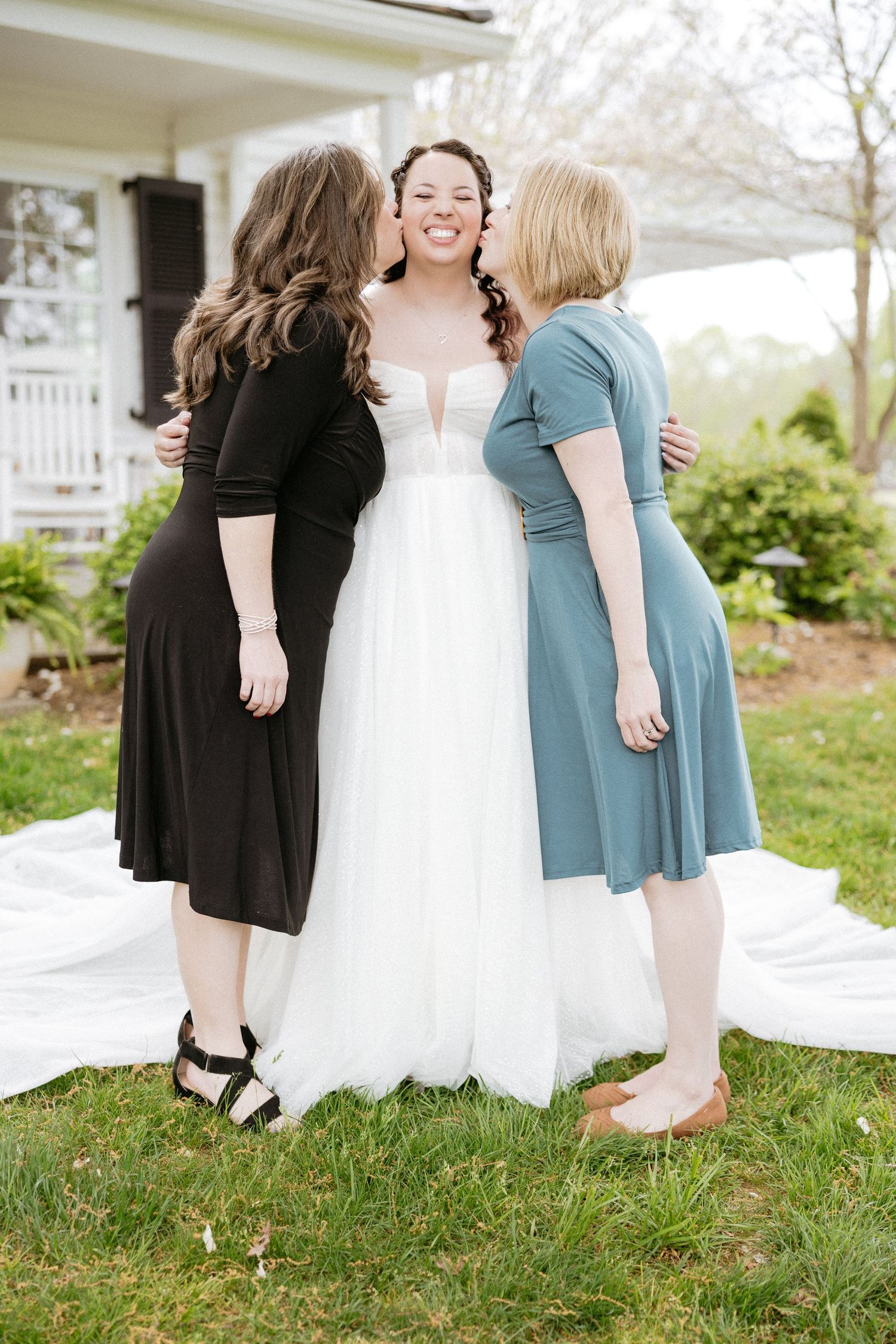 Bride in white gown poses with friends wearing black and blue dresses on grassy lawn.