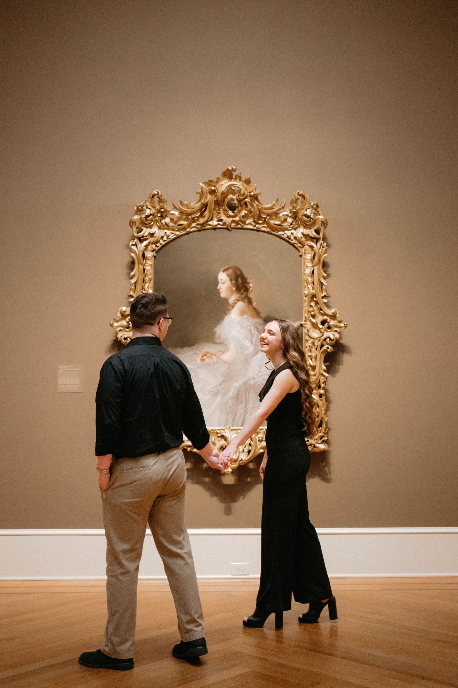Two visitors engage with an ornate gold-framed mirror in an art gallery with wood floors and beige walls.