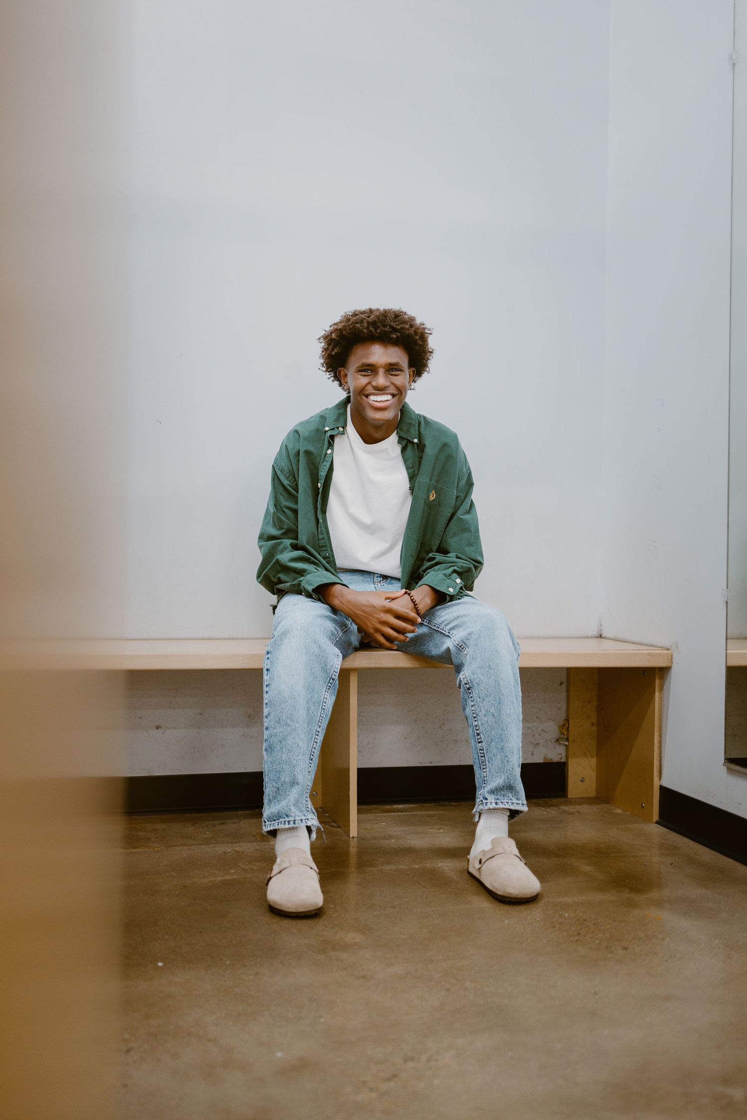 Young person in green jacket and jeans sits casually on wooden bench against gray wall while smiling.
