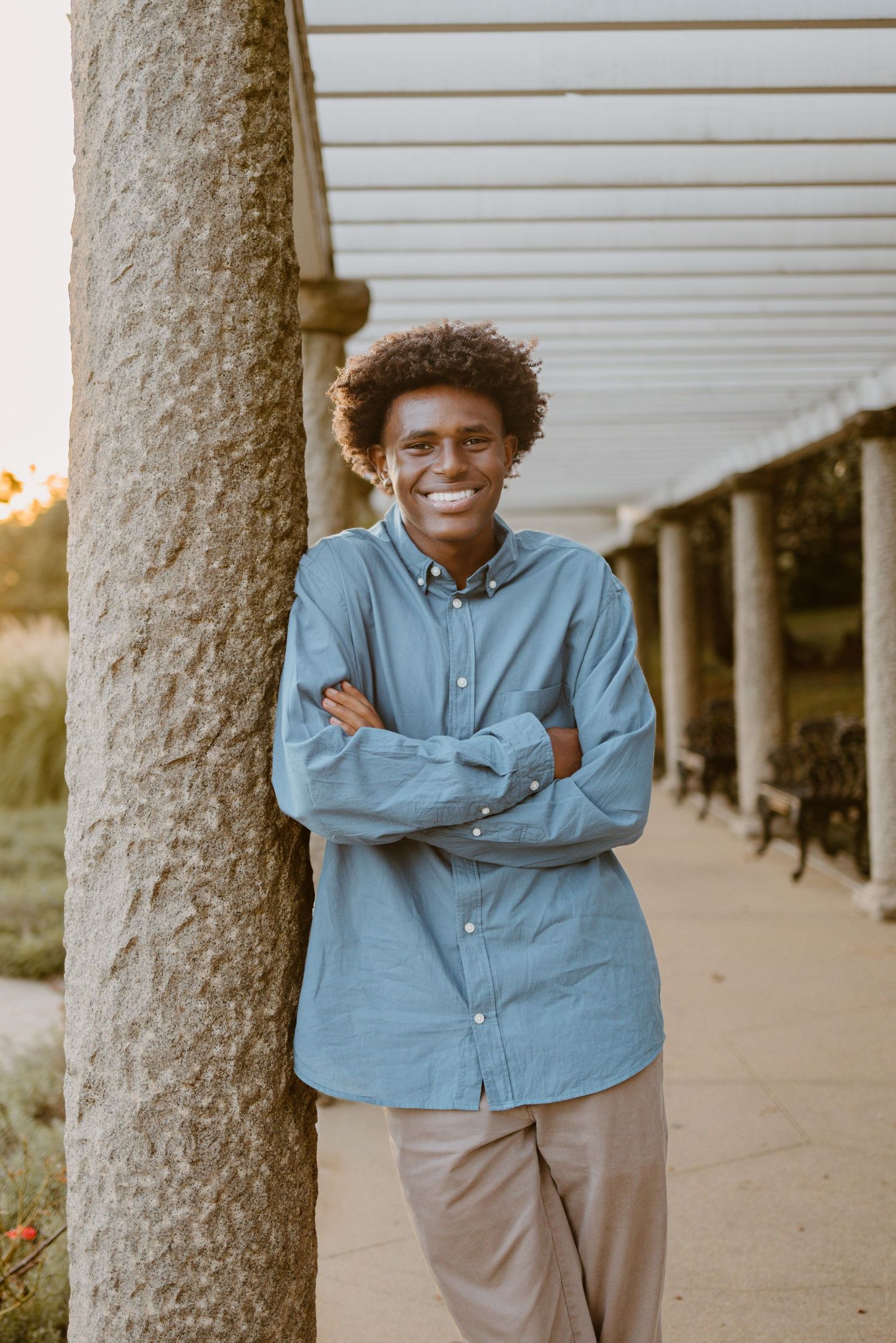 Person in blue button down shirt leans against concrete pillar outdoors at sunset while smiling and enjoying the day.