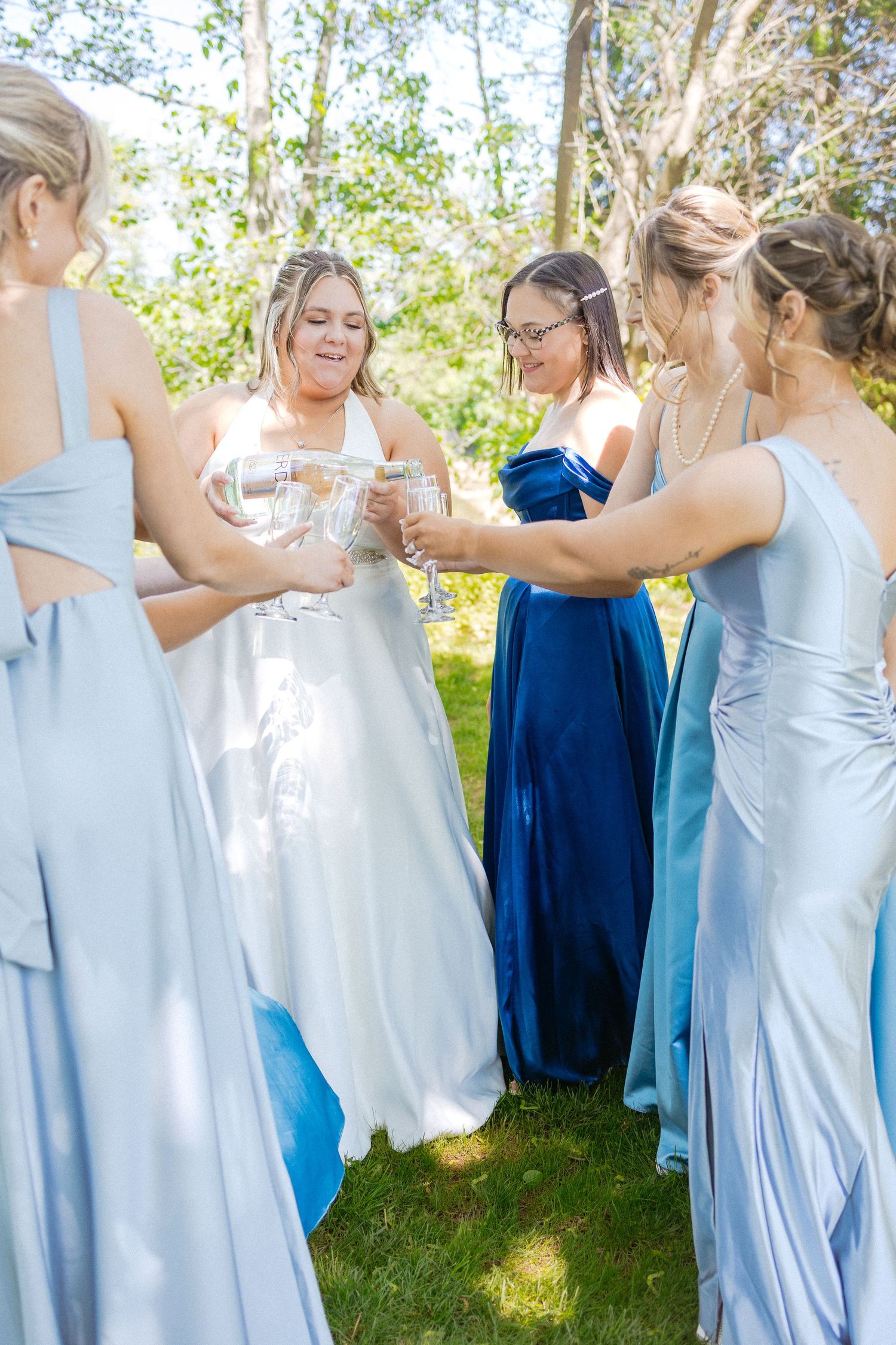 Wedding party of bridesmaids in shades of blue and light colored dresses gather outdoors on a sunny day.