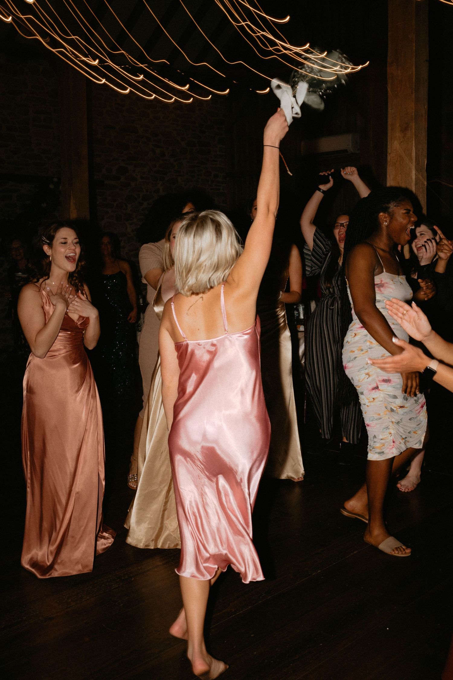 Wedding guests dancing and celebrating at a dimly lit reception with pink and floral dresses visible.
