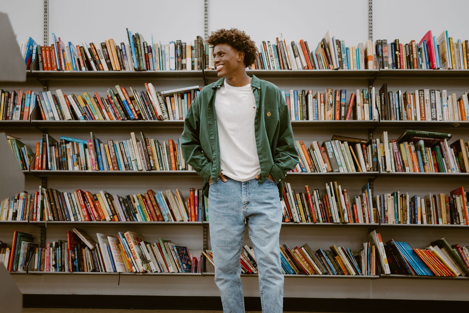 Student standing between rows of library bookshelves with hands casually in pockets.