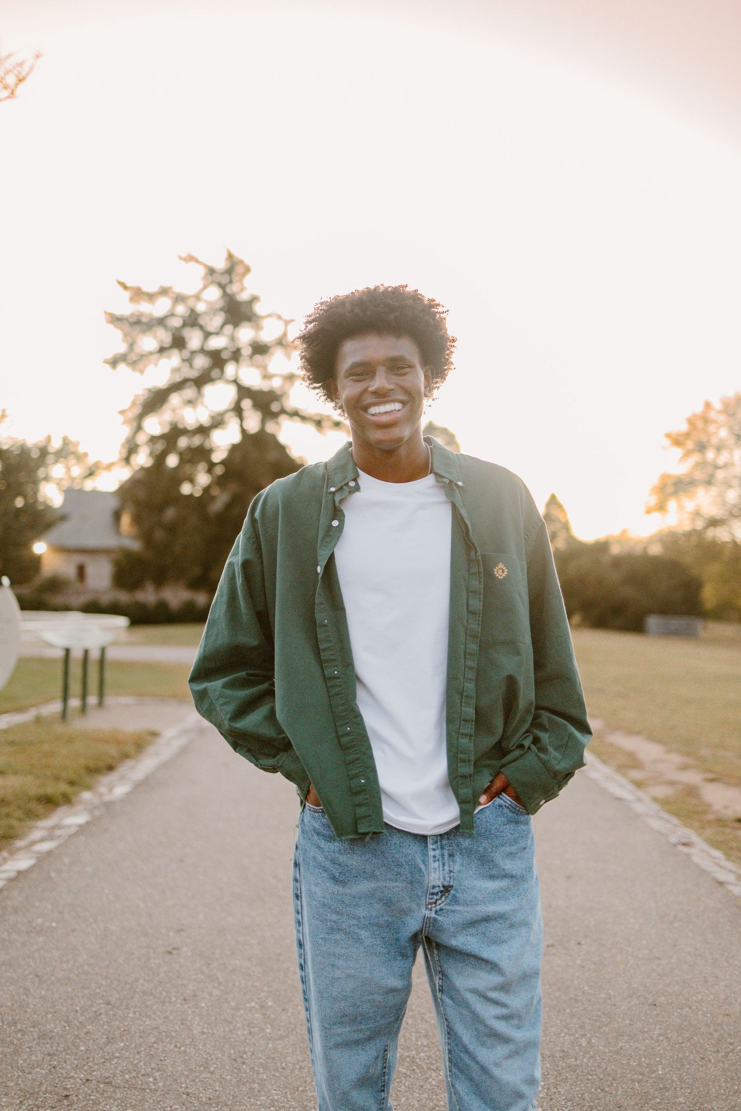 A person in a green jacket and jeans poses with a cheerful expression on a path during sunset.