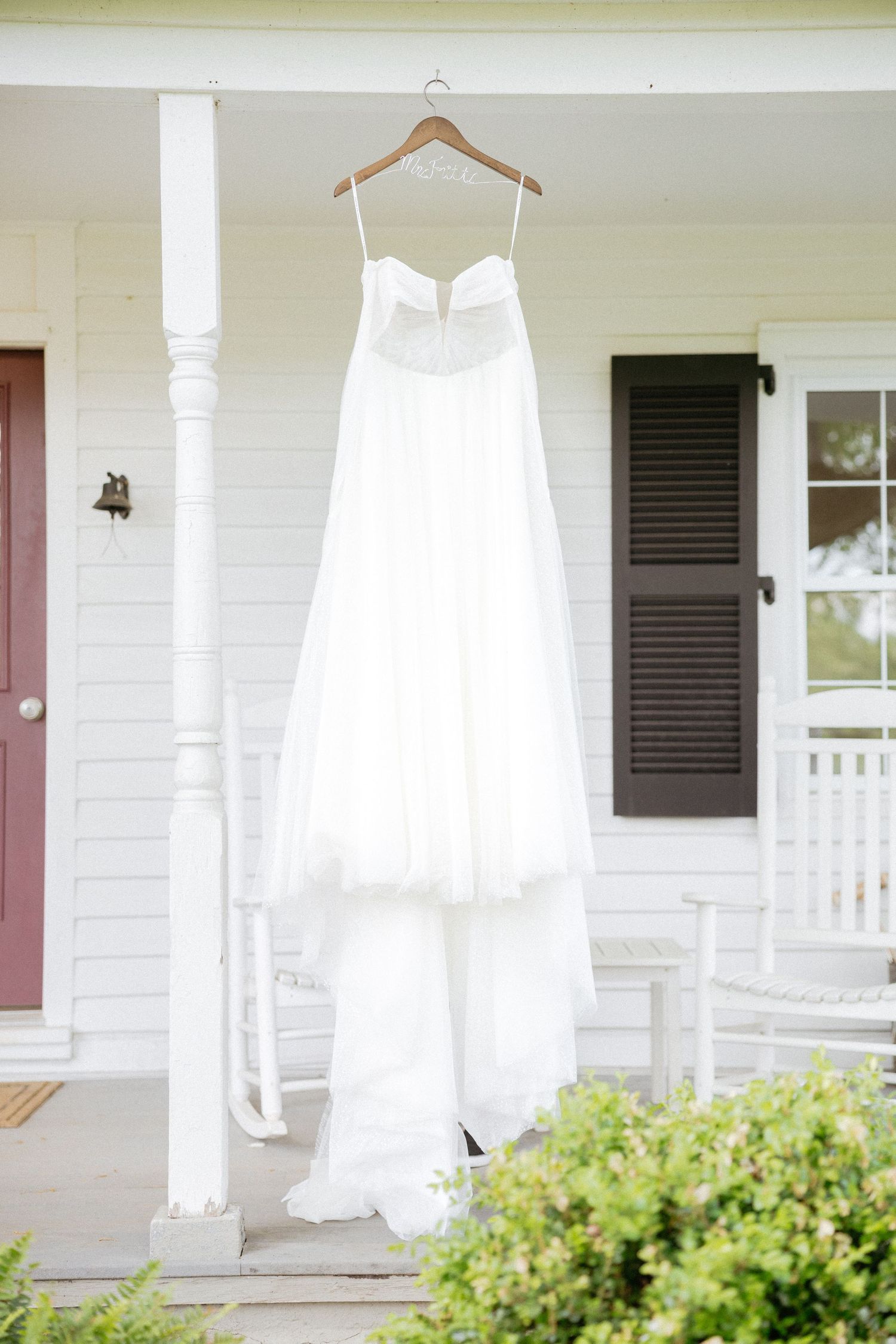 A white wedding dress hangs on a wooden hanger on a porch with green shrubs and white siding.