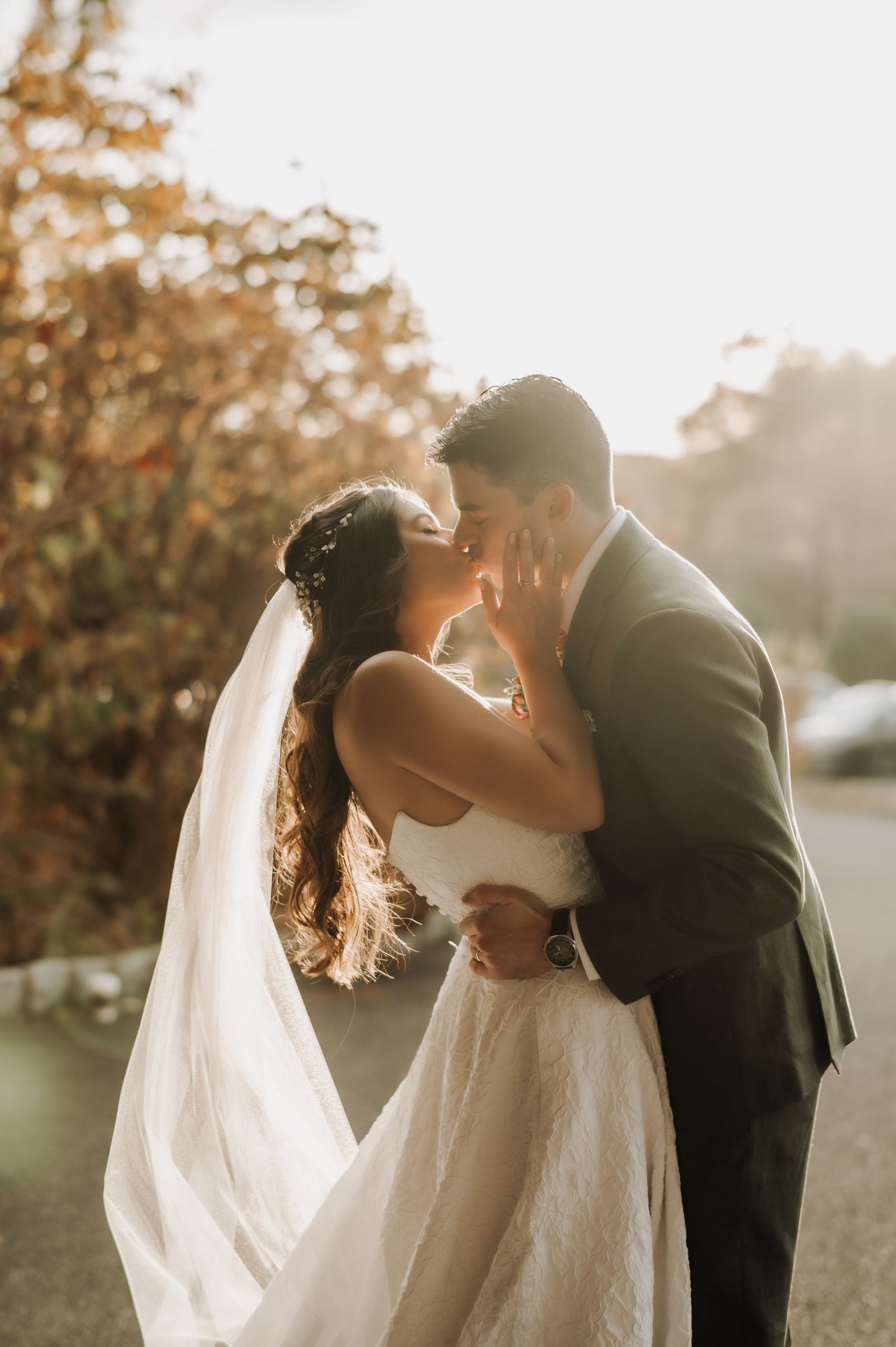 A romantic wedding couple shares an intimate kiss in golden sunlight with a flowing white veil and formal attire.