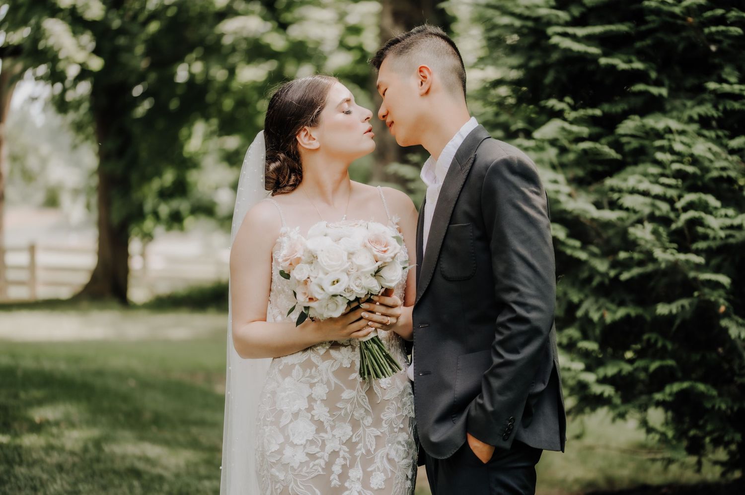A romantic wedding couple embraces outdoors surrounded by lush green trees and natural sunlight.