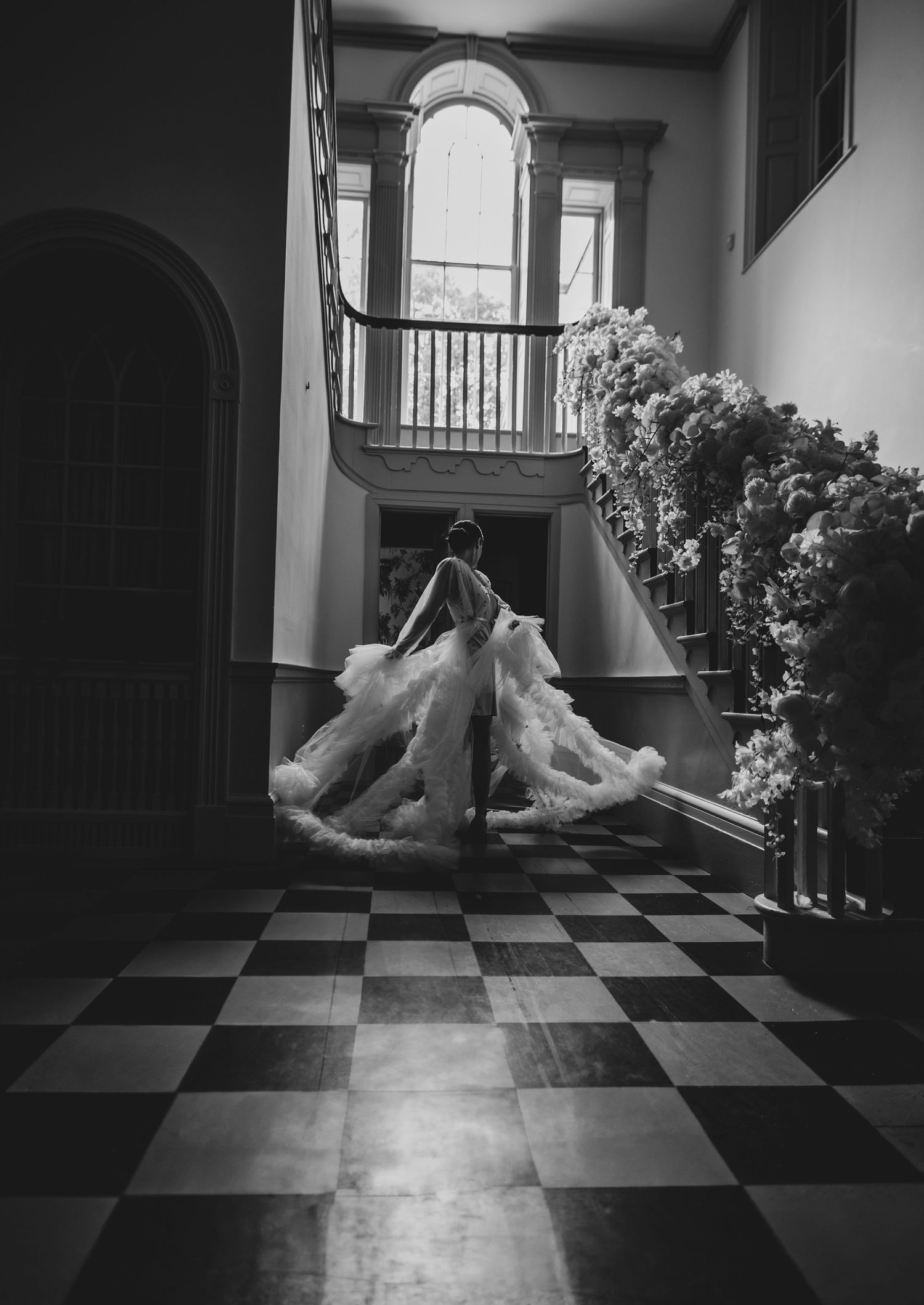 A dramatic black and white photograph of a flowing wedding dress bride on a checkered floor in an elegant interior stairwell.