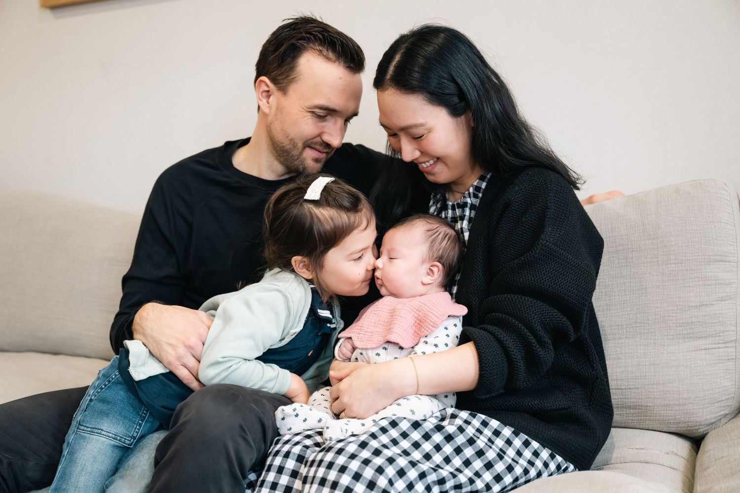 Intimate family moment captured on a light gray couch showing parents cuddling with their newborn baby.