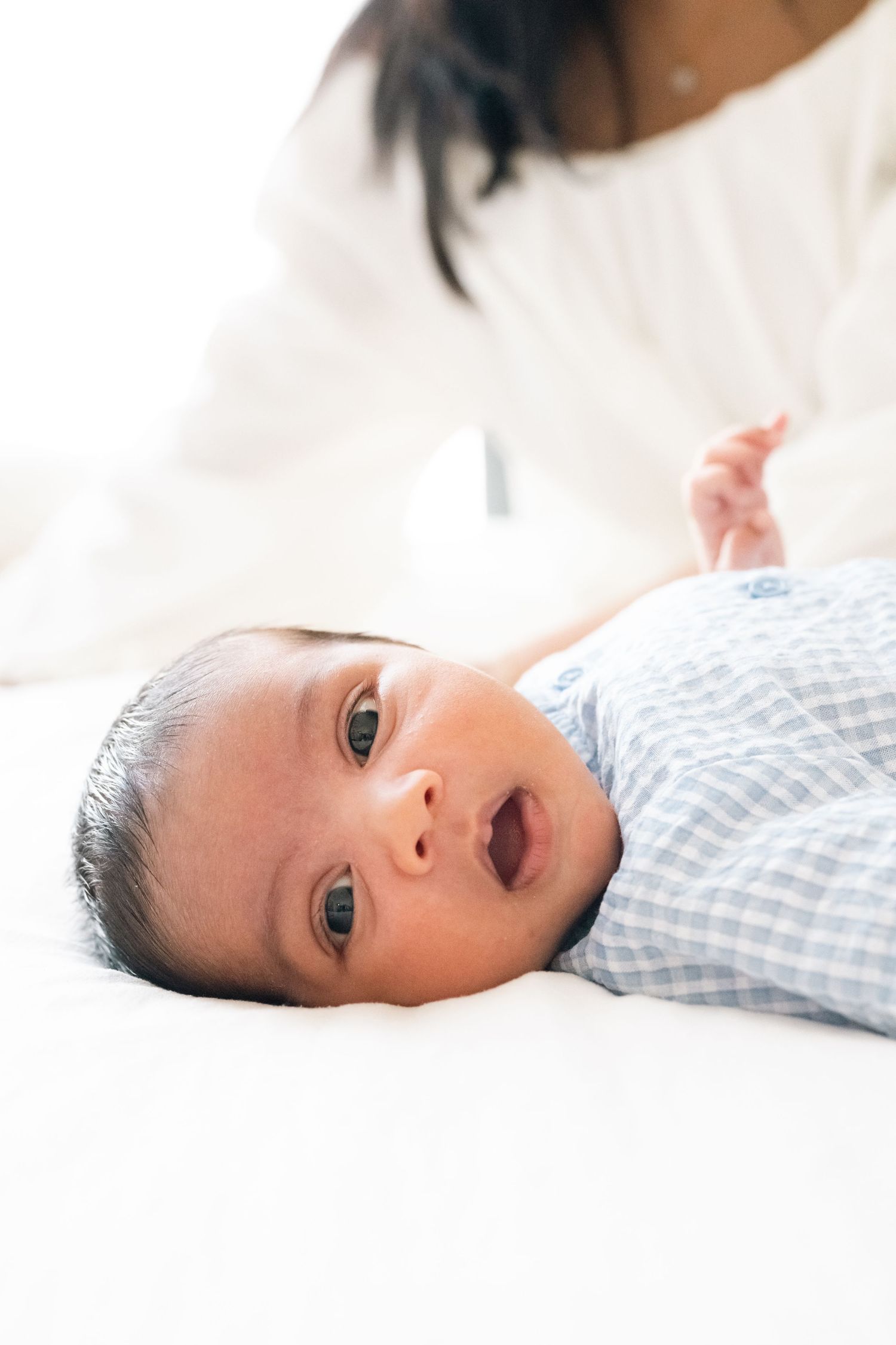 Alert baby in blue checkered outfit gazing up while lying on white bedding.