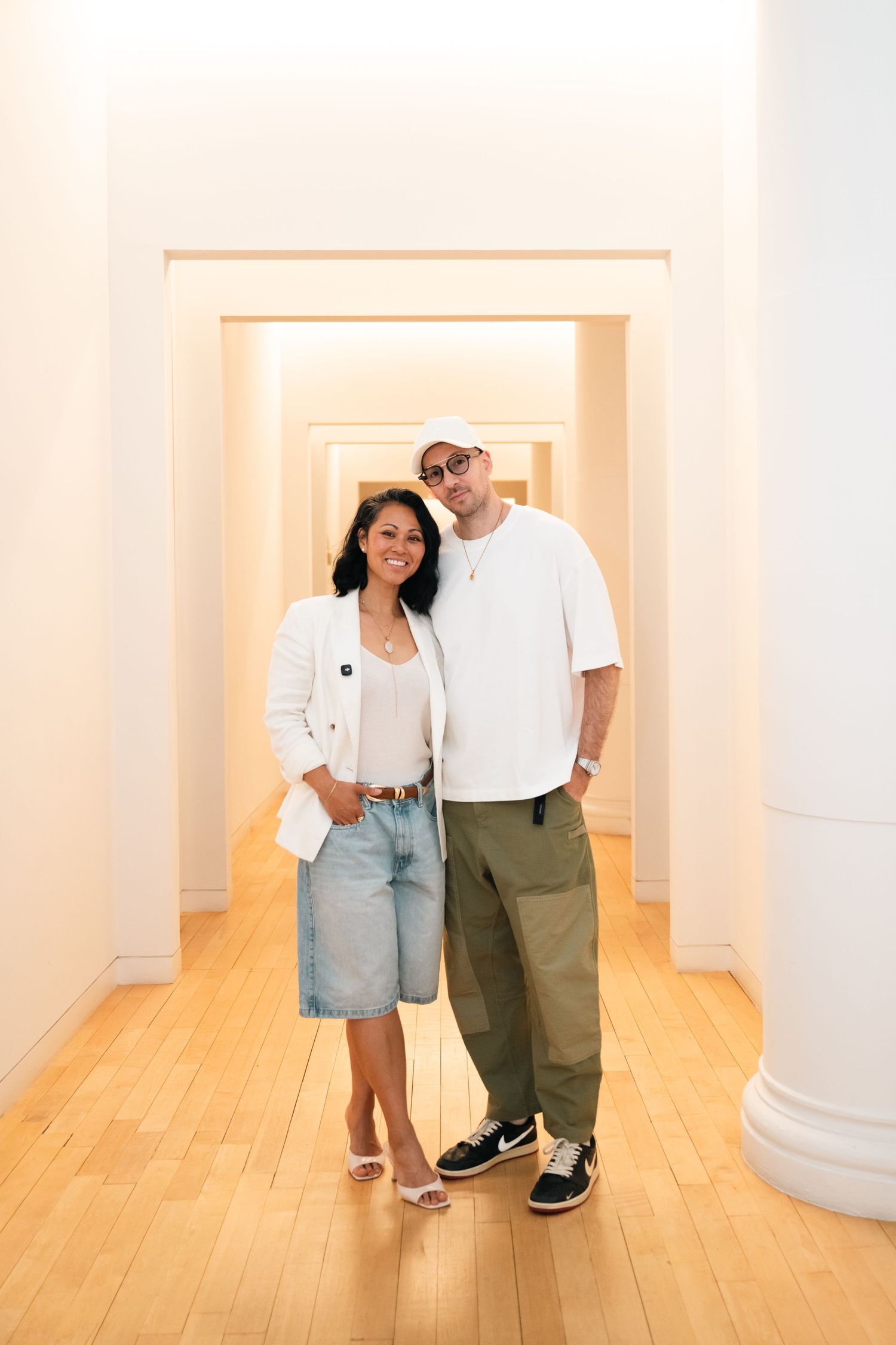 A couple poses together in a bright white hallway with wood floors, wearing casual summer outfits.