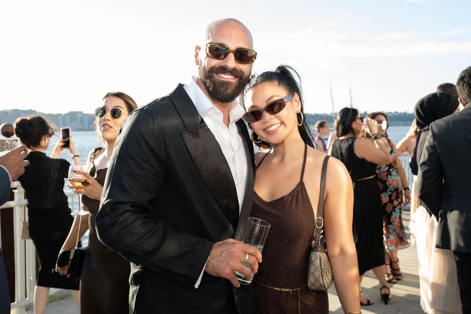 Couple smiling together at an outdoor waterfront event on a sunny evening, man holding a champagne glass.